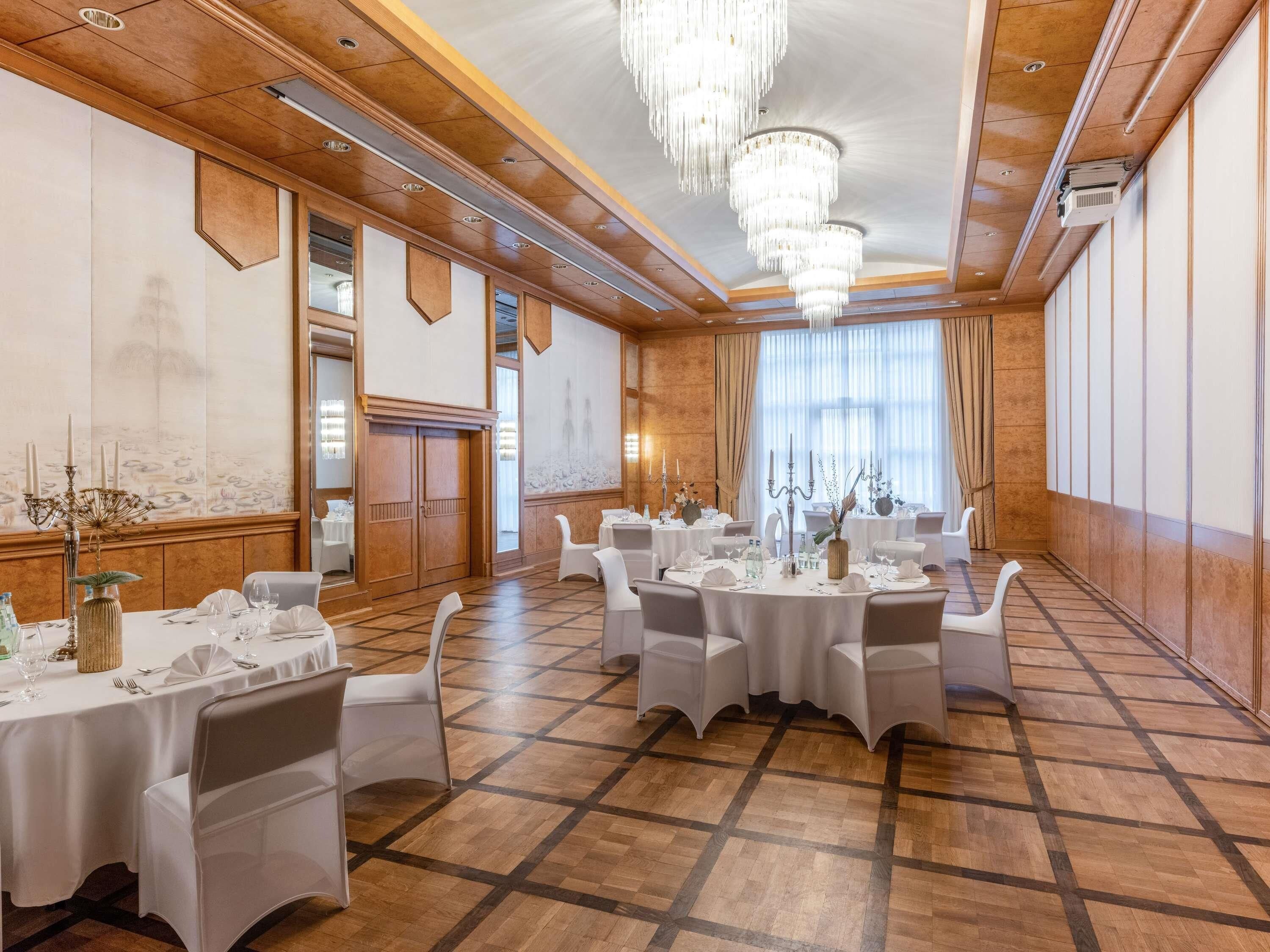 A banquet hall with round tables, white chairs, chandeliers, and a large window with curtains.
