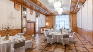 A banquet hall with round tables, white chairs, chandeliers, and a large window with curtains.