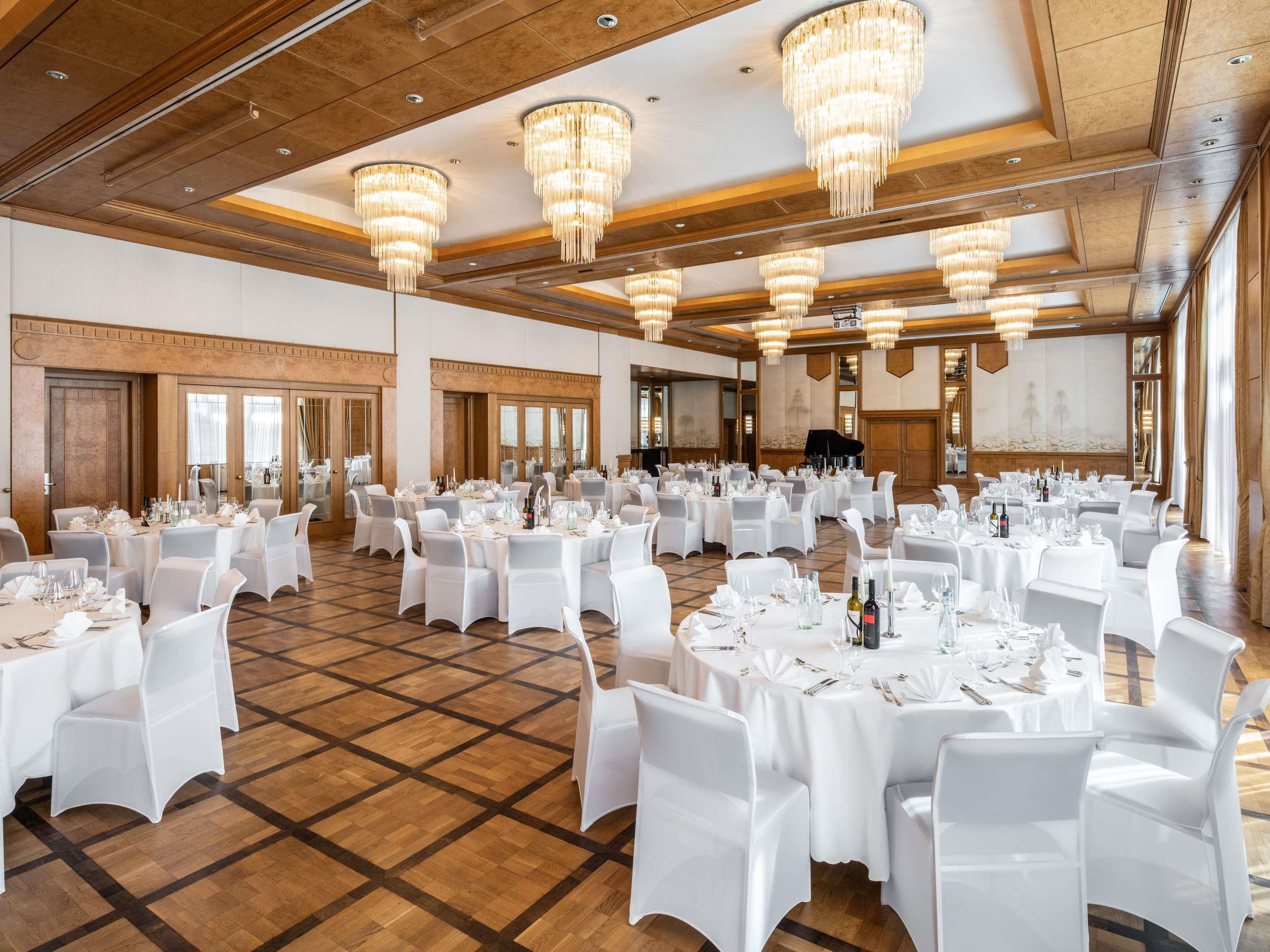 A large banquet hall with round tables set for dining, white chairs, and chandeliers.