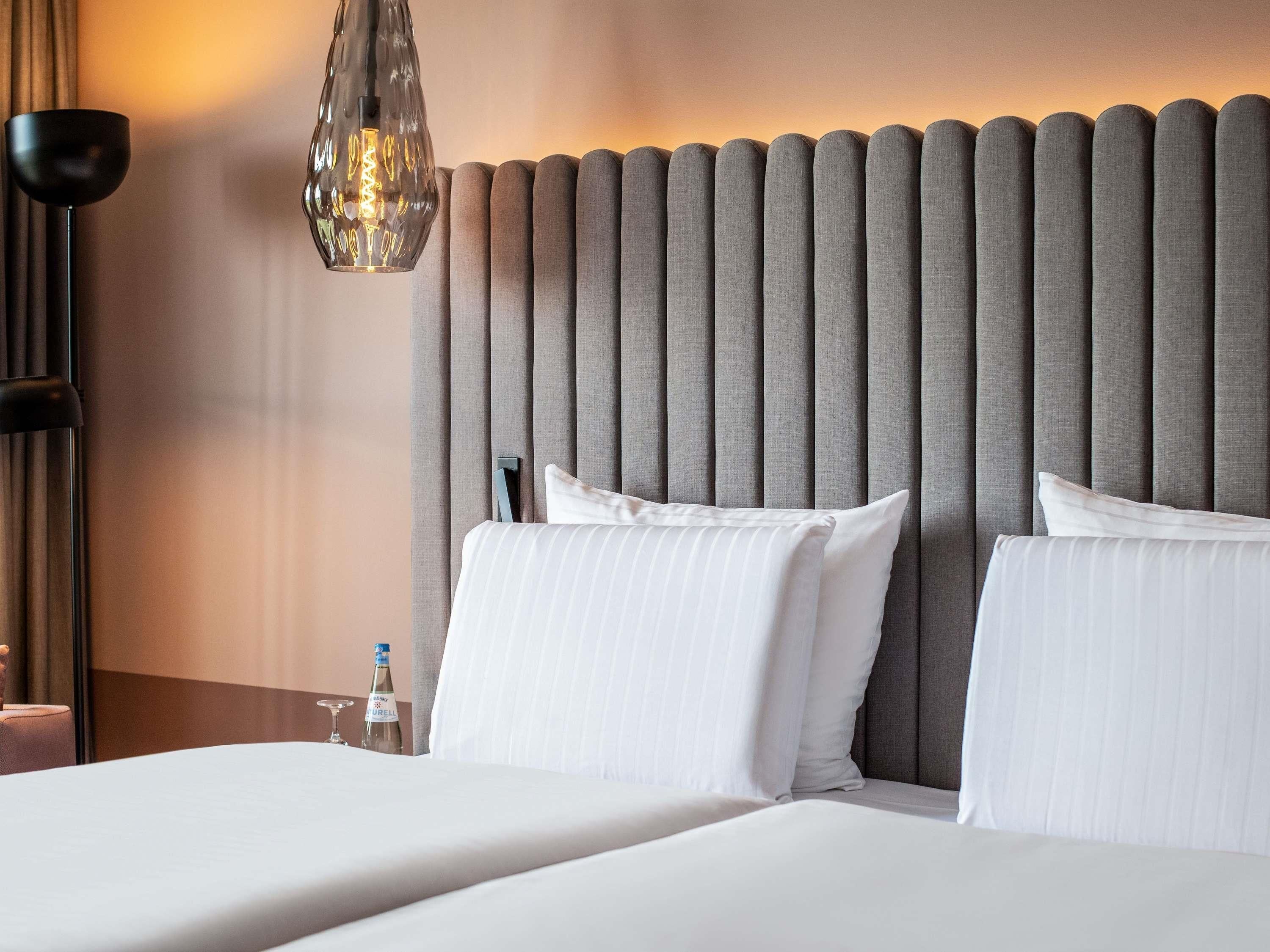 A hotel room with a bed, two pillows, a nightstand, a bottle, and a lamp.