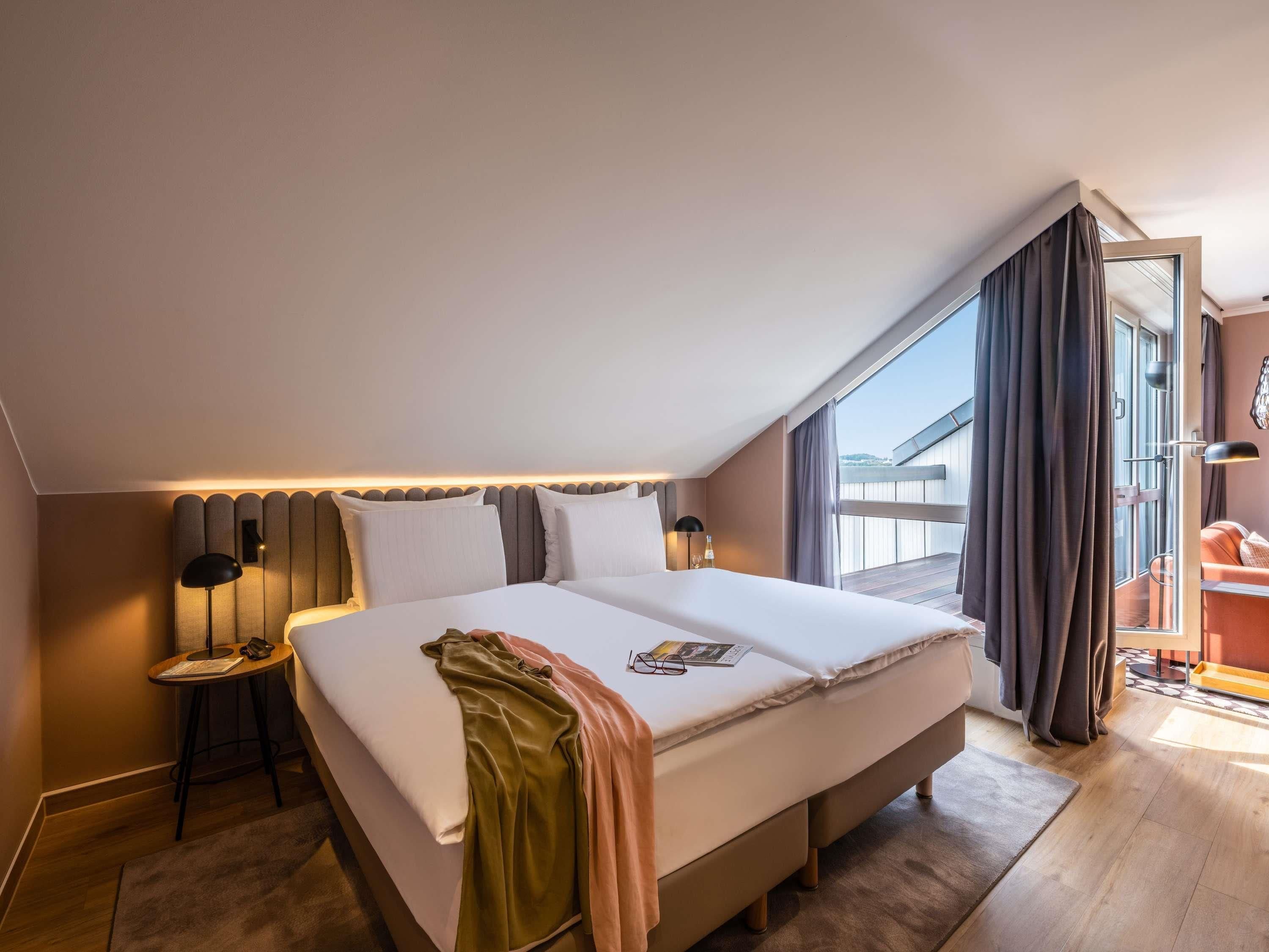 A modern hotel room with a large bed, a side table, a reading lamp, and a view of the outside.