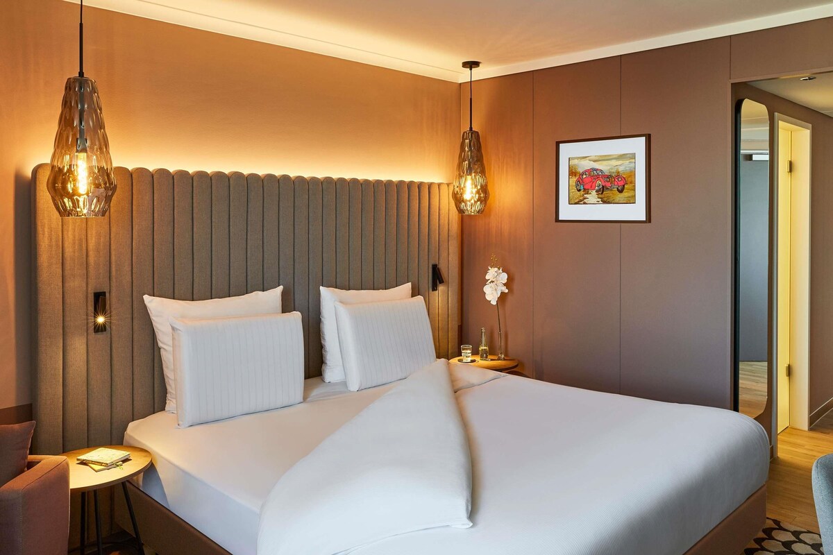 Ein Hotelzimmer mit einem großen Bett, zwei Hängeleuchten, einem kleinen Tisch mit einem Buch und einem gerahmten Bild an der Wand.