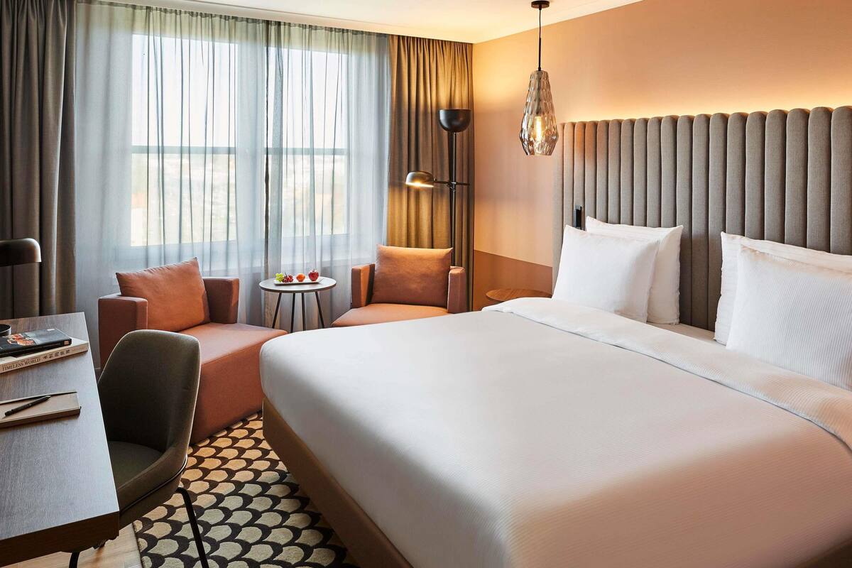 Habitación de hotel con una cama grande, un escritorio, una silla, una mesita con fruta y una lámpara.