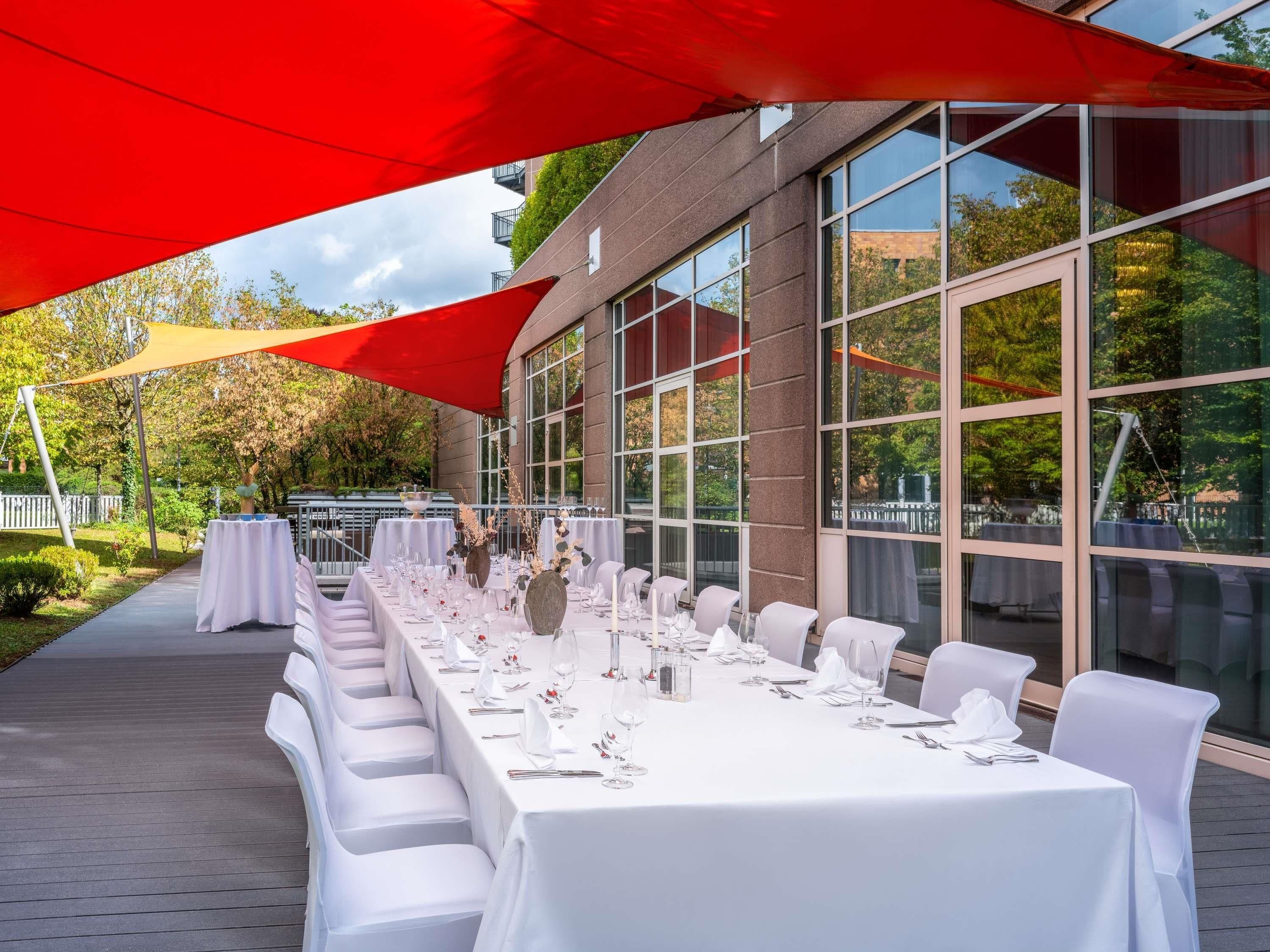 Un área de comedor al aire libre con mesas dispuestas para una comida, amplias ventanas y un edificio de ladrillo al fondo.