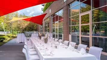 Un área de comedor al aire libre con mesas dispuestas para una comida, amplias ventanas y un edificio de ladrillo al fondo.
