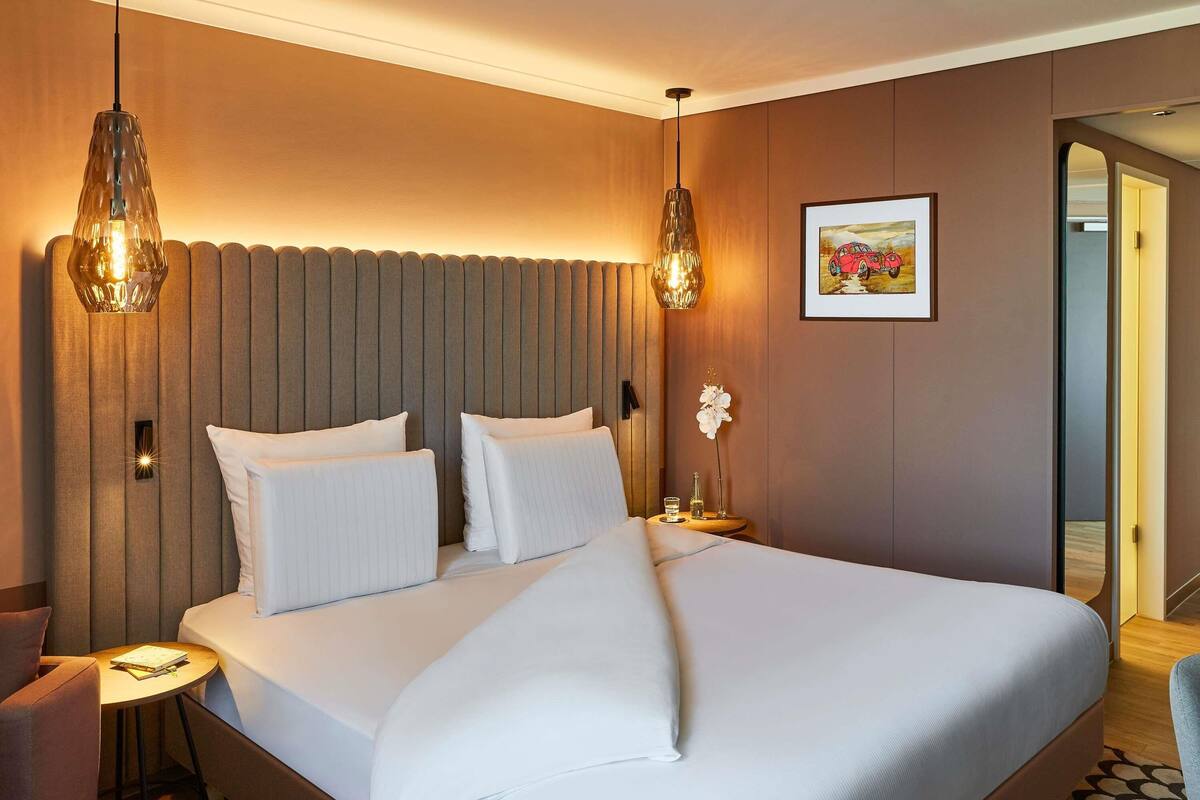 Habitación de hotel con una cama grande, dos lámparas colgantes, una mesita con un libro y un cuadro en la pared.
