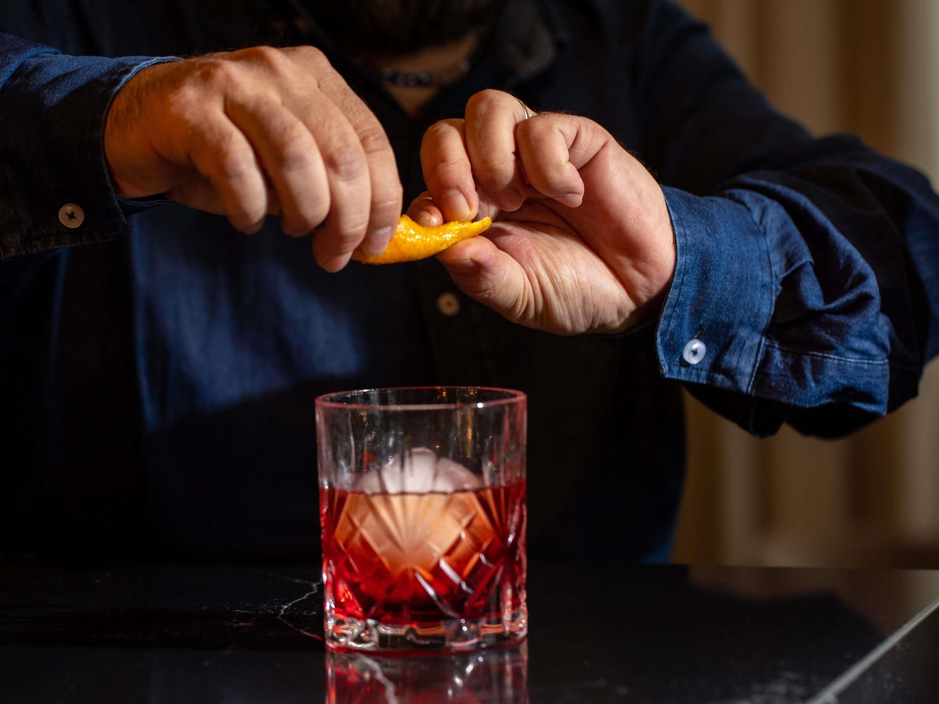 En person presser en citron ud over et glas rød væske, sandsynligvis en cocktail, på en mørk overflade.