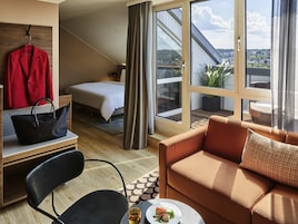 A hotel room with a bed, a sofa, a chair, a table with a plate and glass, a hanging coat, and a large window with a view.