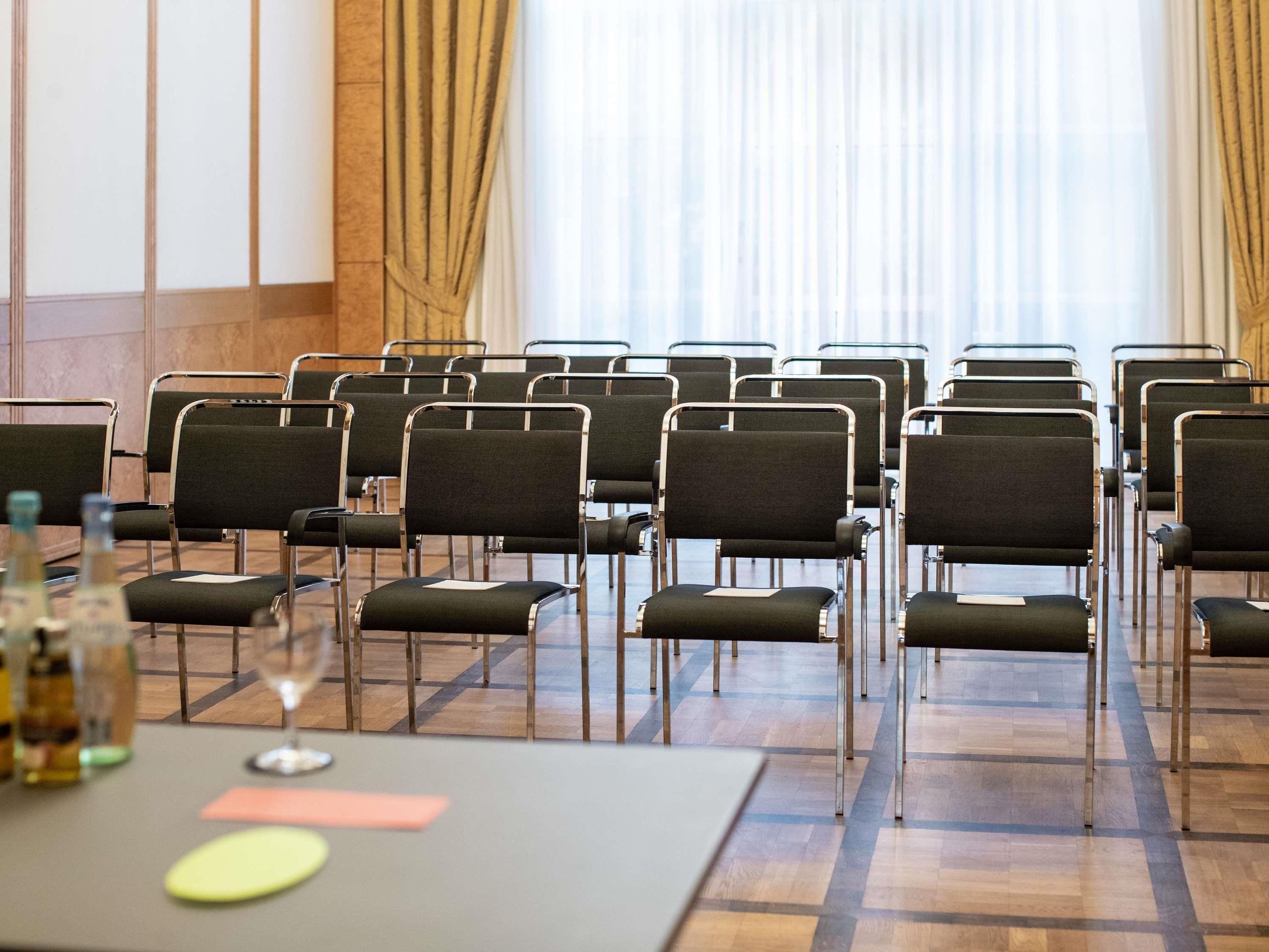 A conference room with rows of chairs, a table with a bottle and glass, and a window with curtains.