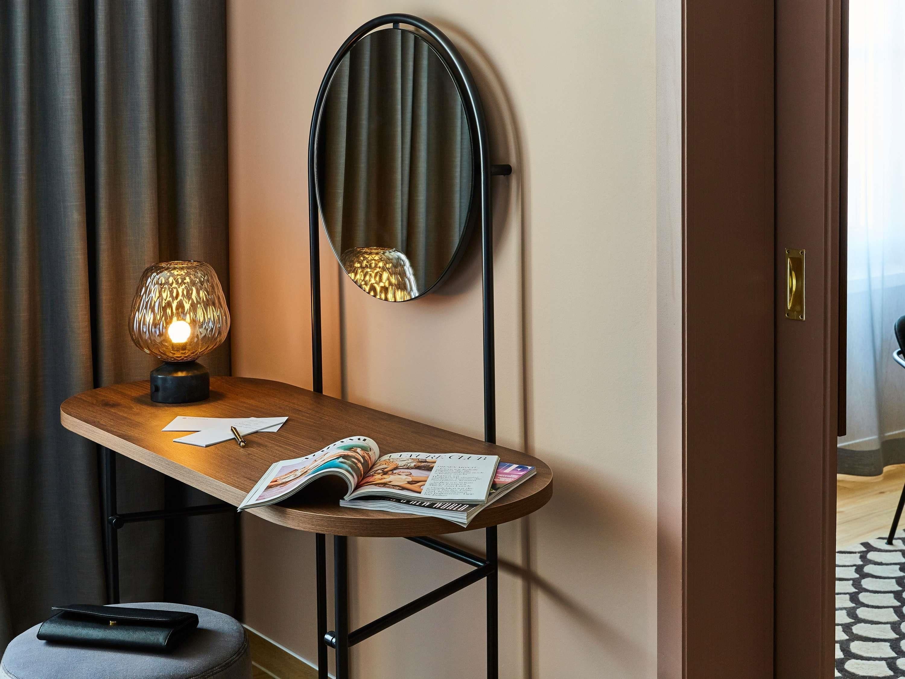 A hotel room with a wooden desk, a round mirror, a lamp, and a magazine.
