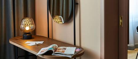 A hotel room with a wooden desk, a round mirror, a lamp, and a magazine.