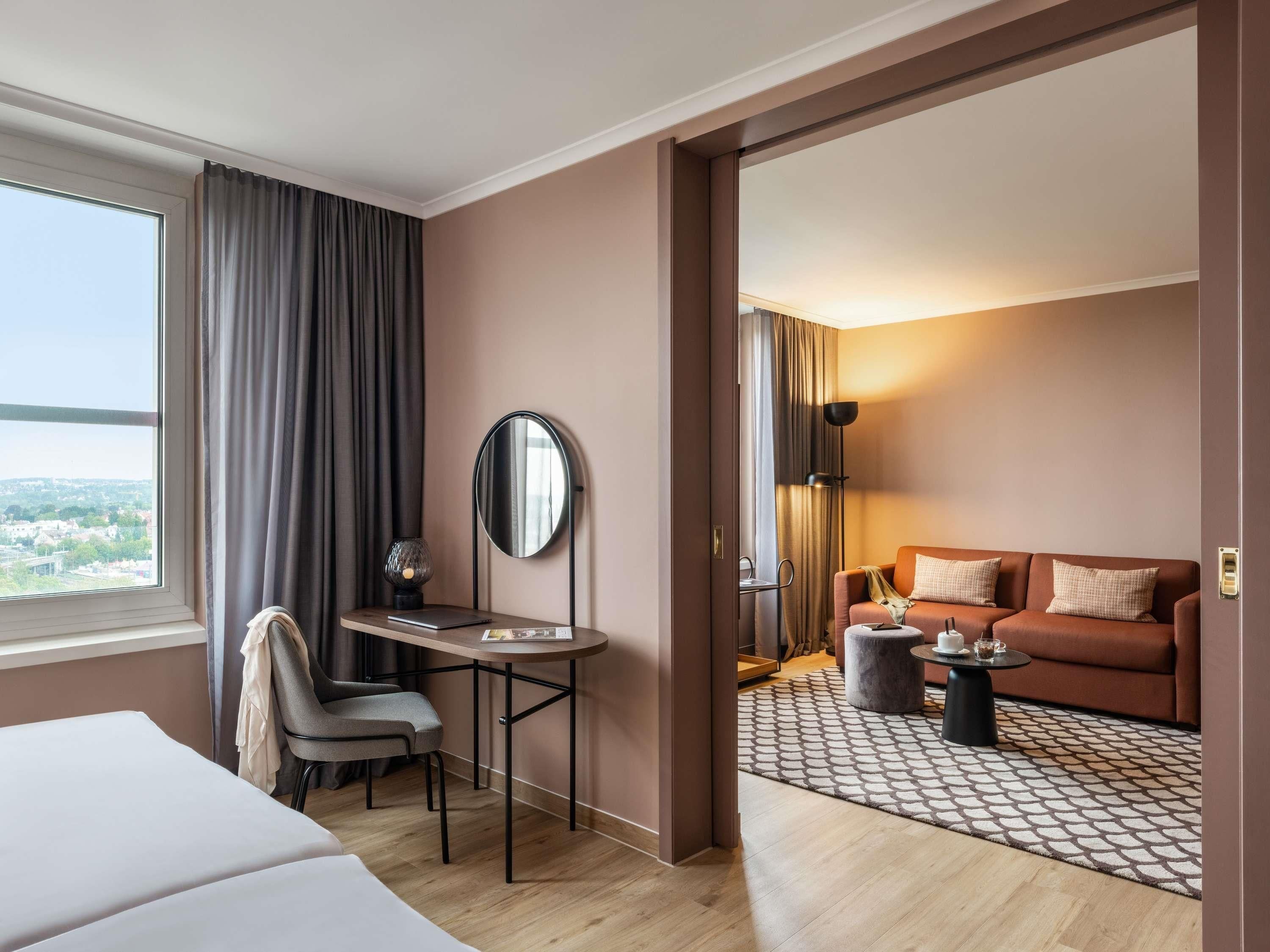Una habitación de hotel con una cama, un escritorio con una silla, un sofá, una mesa de centro y un amplio ventanal con cortinas.