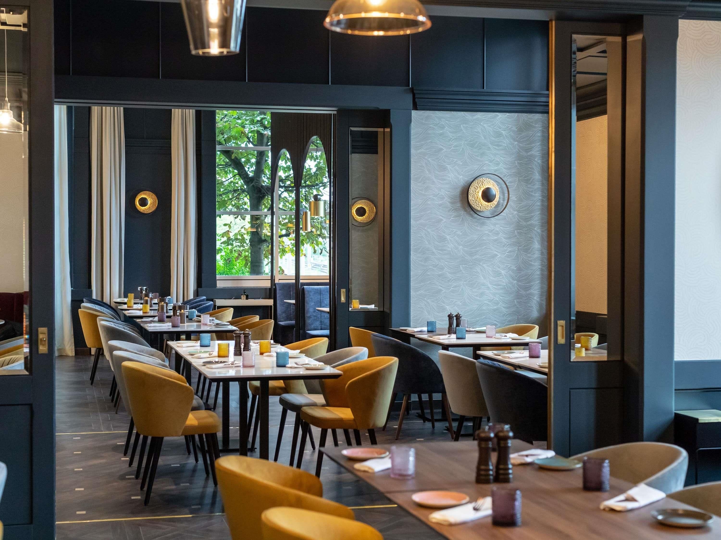 Un restaurante moderno con largas mesas compartidas, sillas amarillas y grandes ventanales con vistas a zonas verdes.