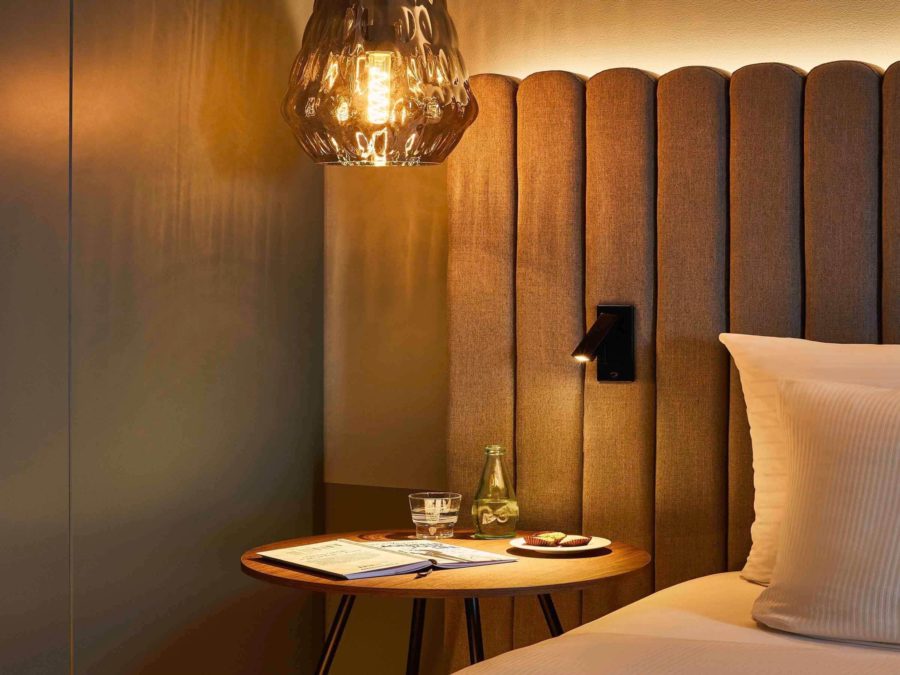 Hotelkamer met een bed, nachtkastje met lamp, wandlamp en een kleine tafel met een glas water en een boek.