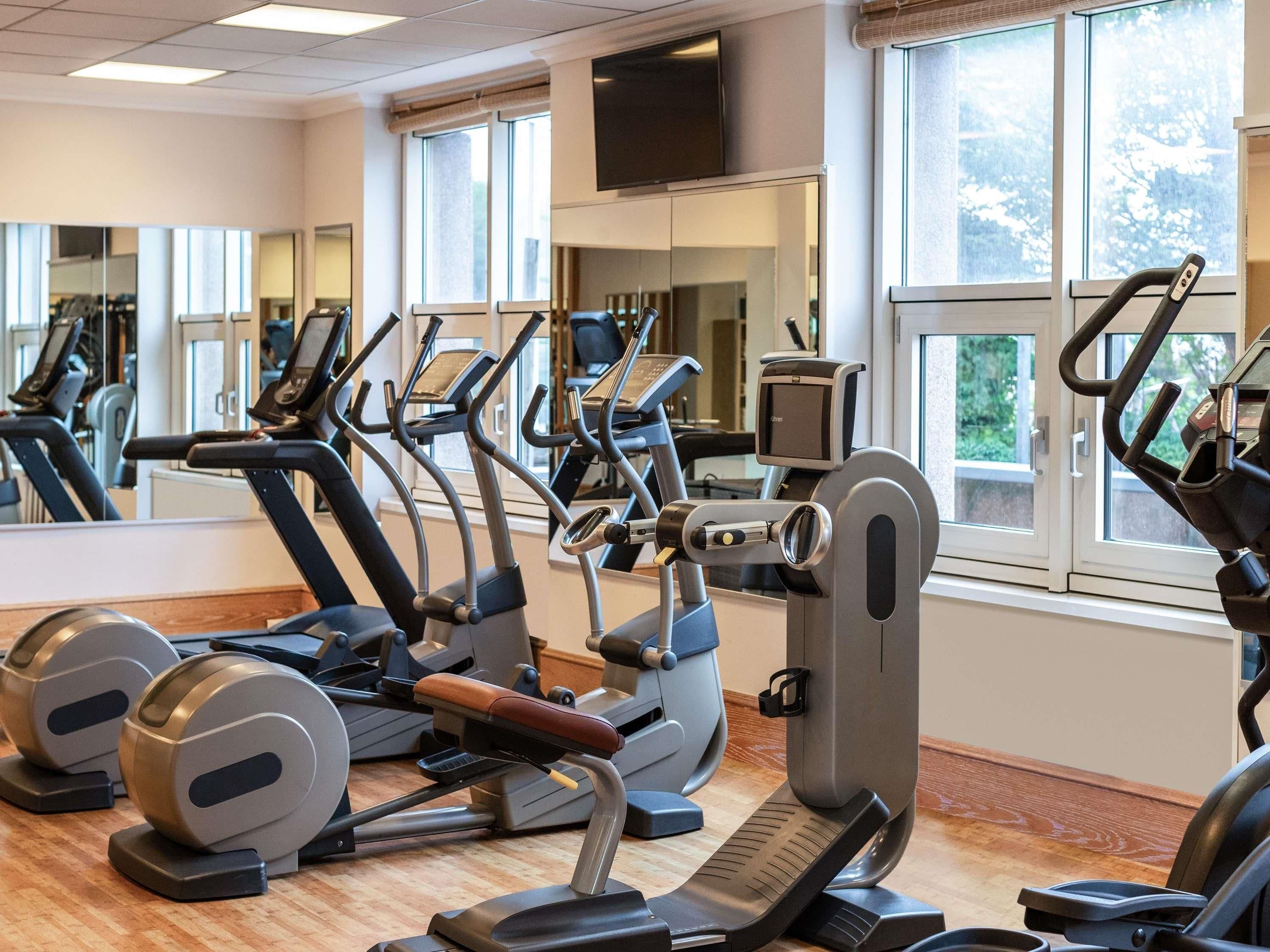 Une salle de sport équipée de divers appareils d’exercice, dotée de grandes fenêtres et d’un miroir.