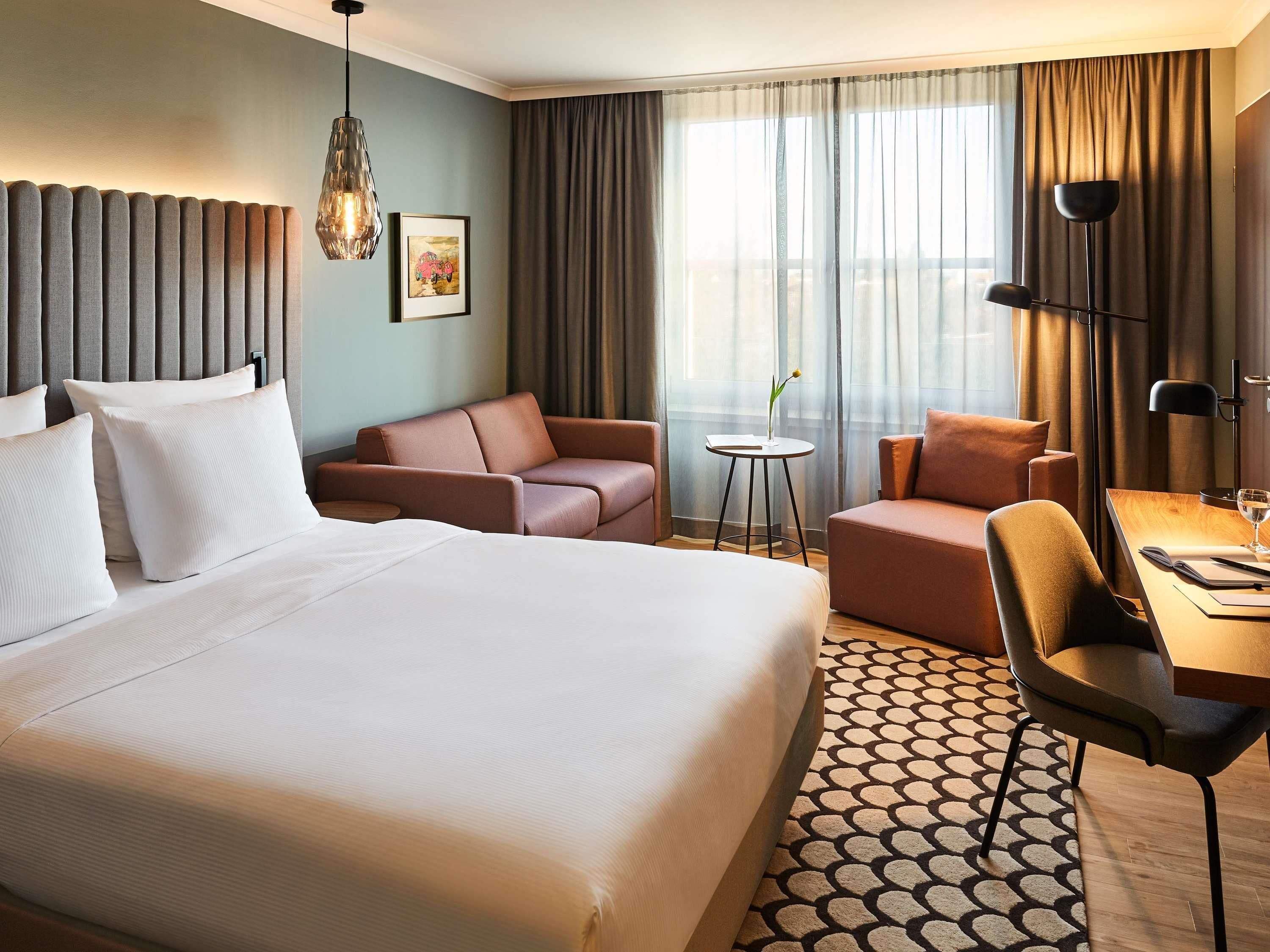 A hotel room with a large bed, two sofas, a small table with a lamp, and a view of the city through the window.
