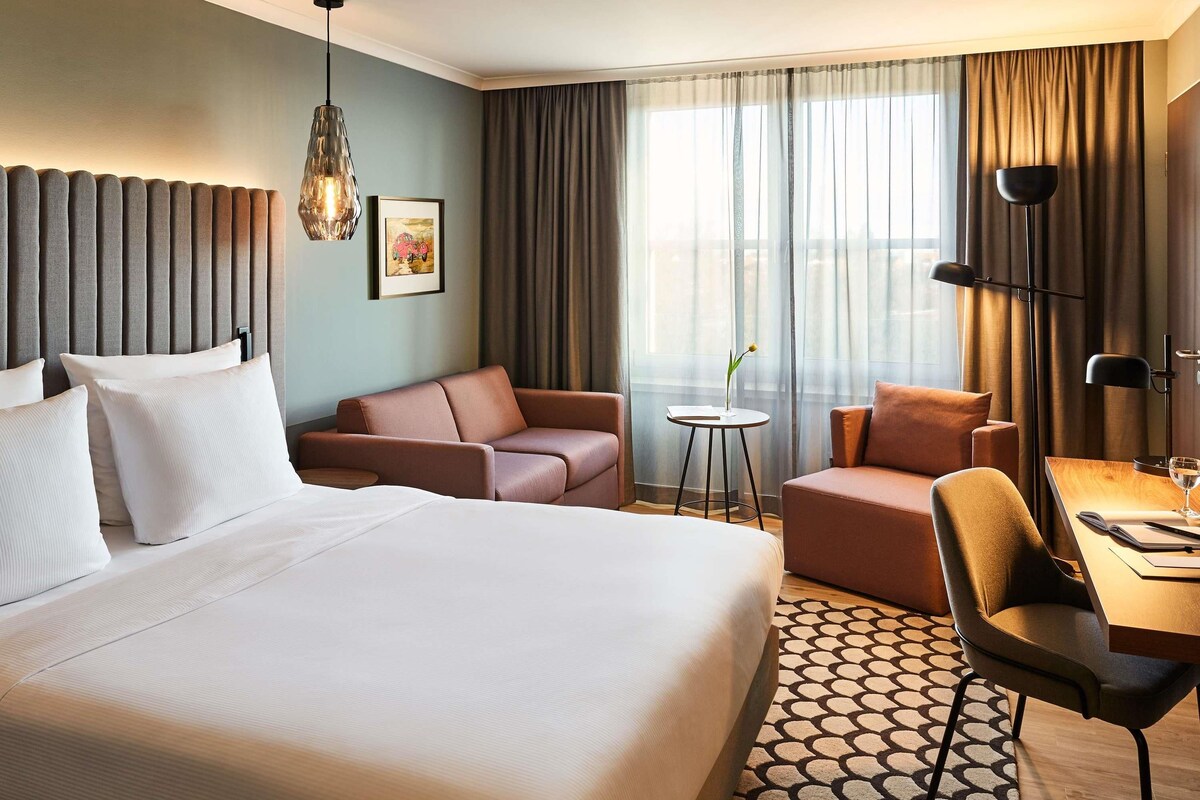 Ein Hotelzimmer mit einem großen Bett, zwei Sofas, einem kleinen Tisch mit Lampe und Blick auf die Stadt durch das Fenster.