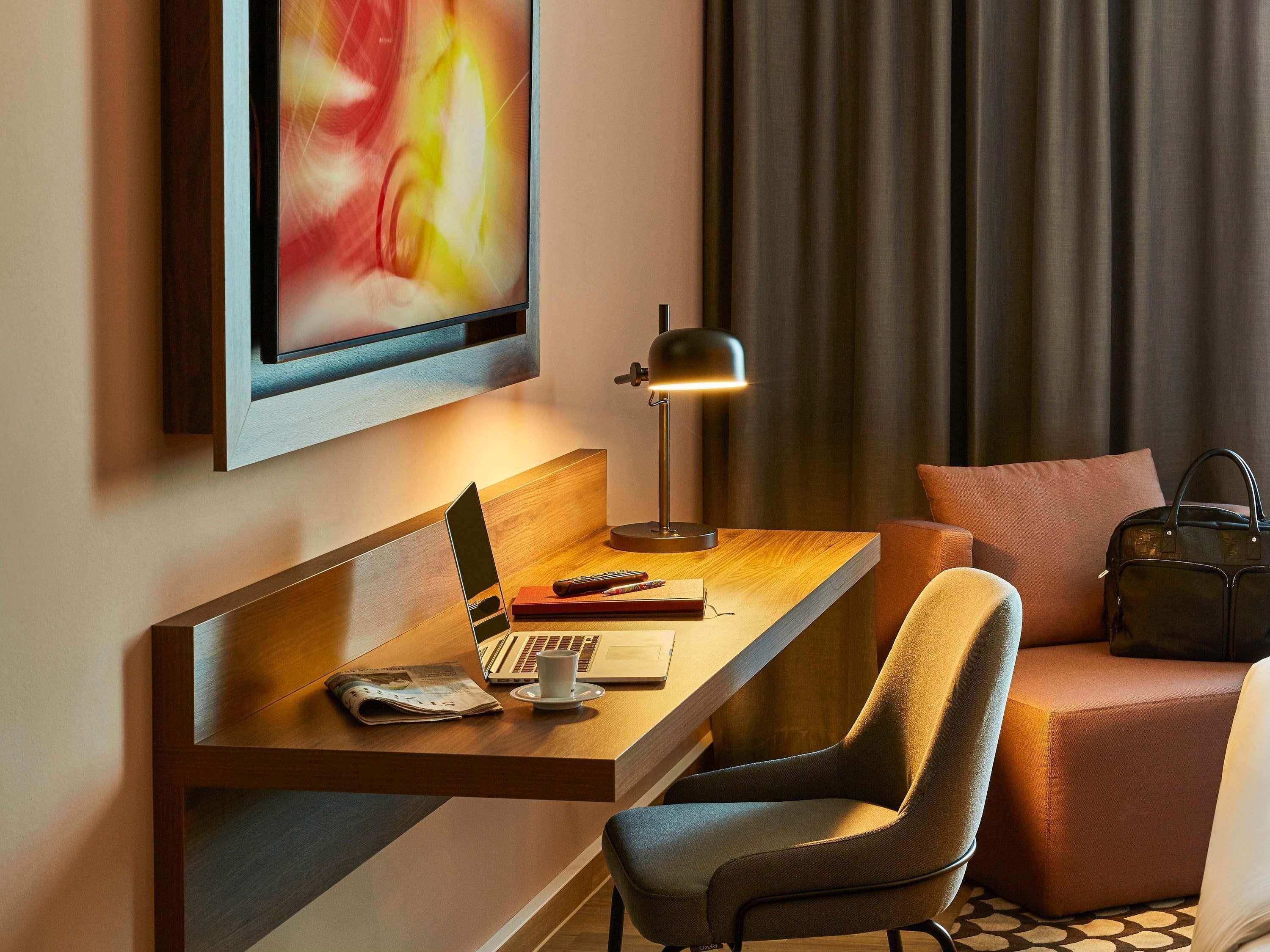 A modern hotel room with a wooden desk, a laptop, a lamp, a chair, a sofa, and a television mounted on the wall.