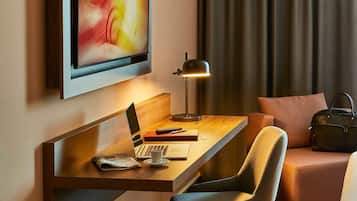 A modern hotel room with a wooden desk, a laptop, a lamp, a chair, a sofa, and a television mounted on the wall.