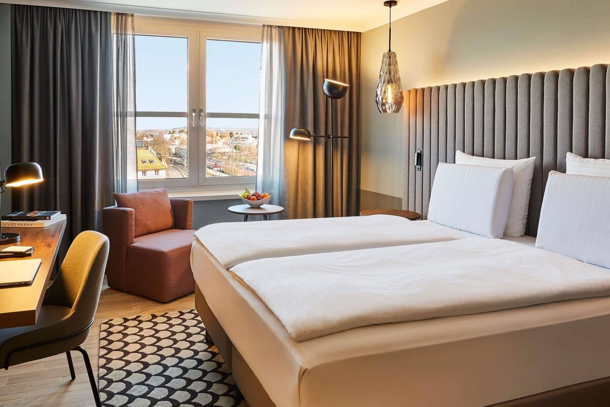 Habitación de hotel con una cama grande, un escritorio con silla, una mesita con frutero, y vistas a la ciudad a través de la ventana.