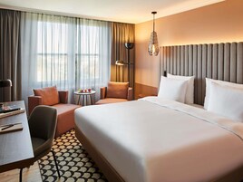 A hotel room with a large bed, a desk, a chair, a small table with fruit, and a lamp.