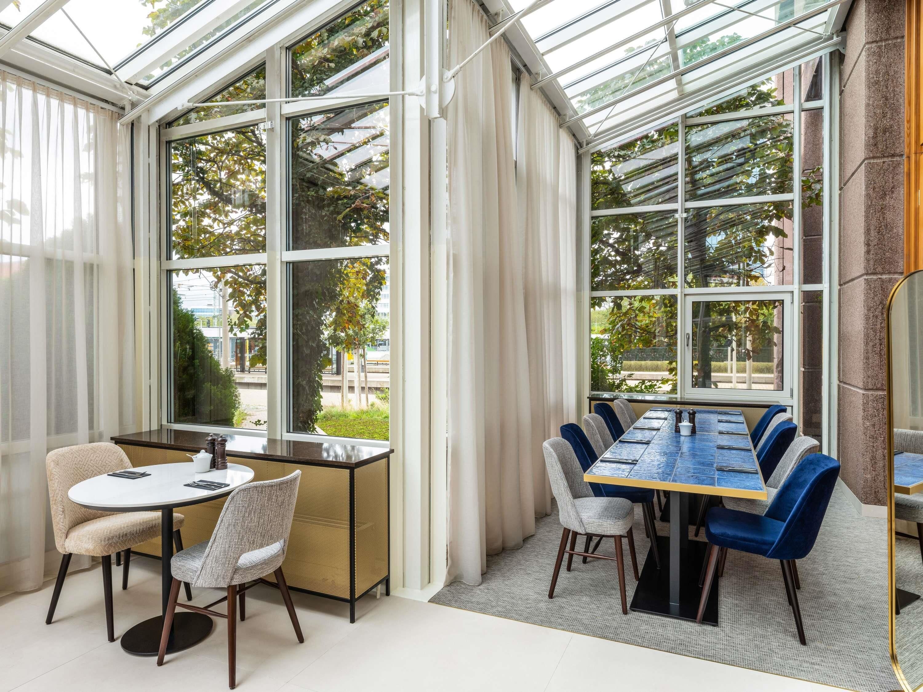 A conservatory with a long table and chairs, large windows, and a view of greenery outside.