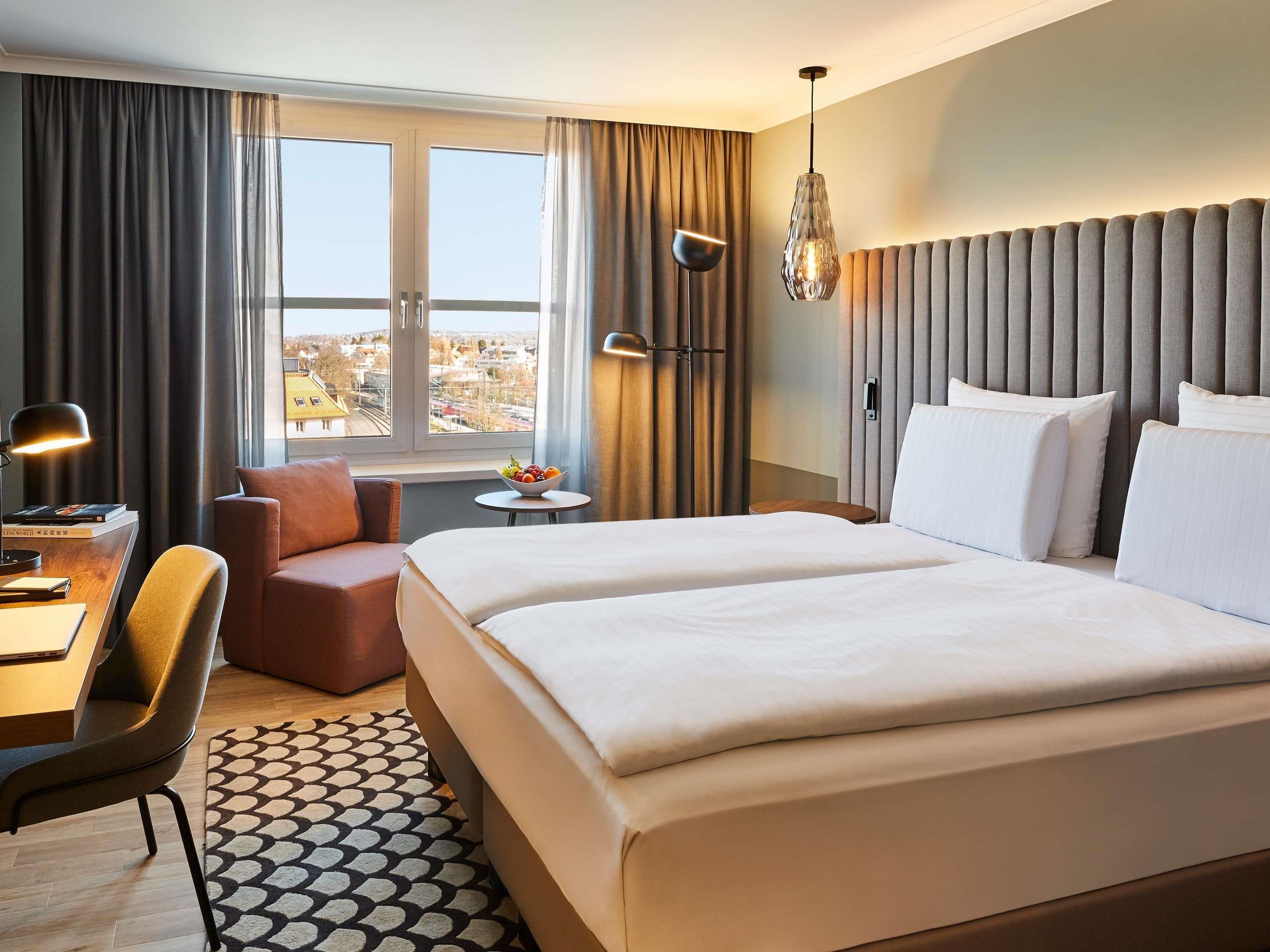 Ein Hotelzimmer mit einem großen Bett, einem Schreibtisch mit Stuhl, einem kleinen Tisch mit Obstschale und Blick auf die Stadt durch das Fenster.