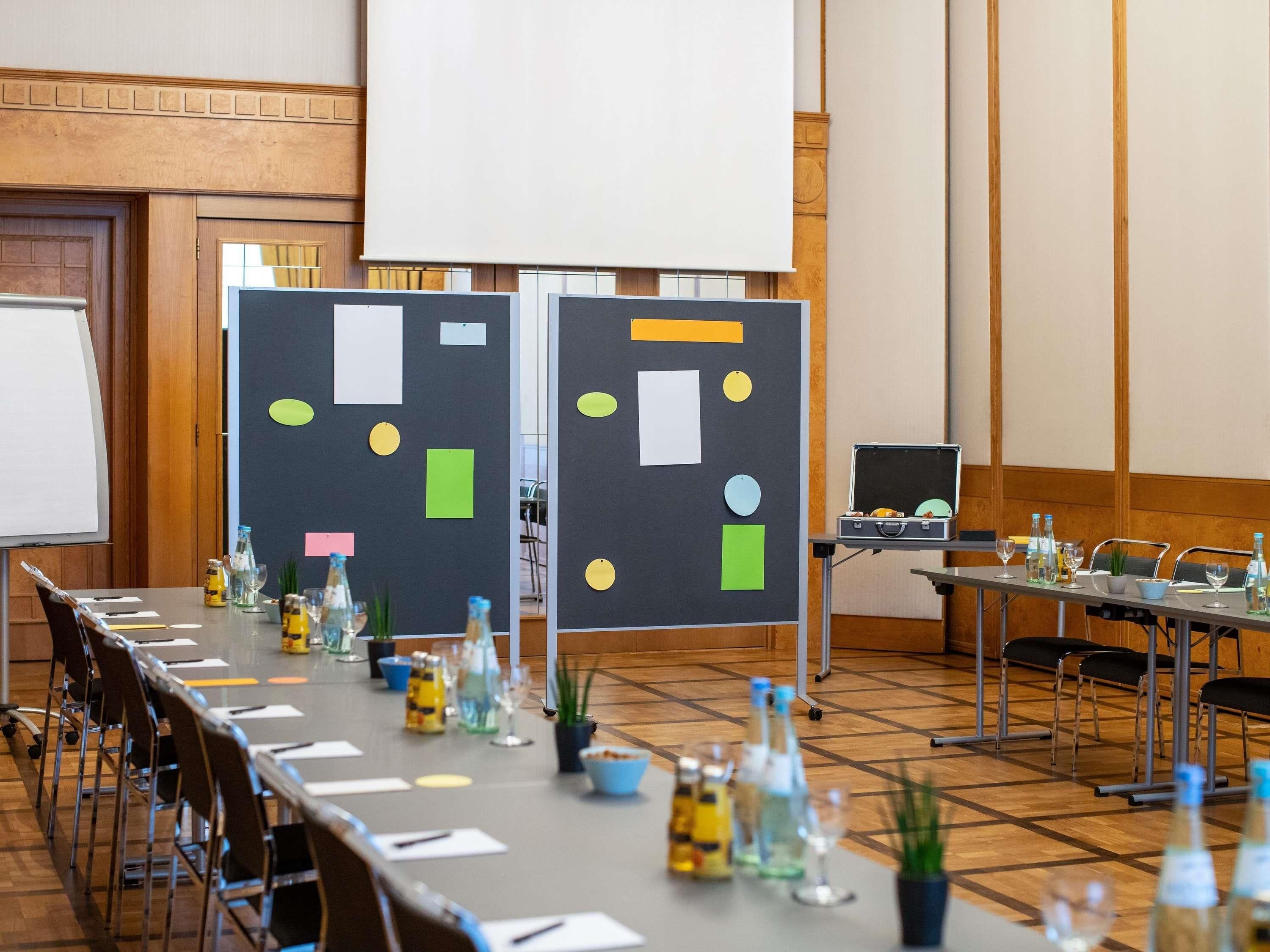 Un salón de conferencias con una larga mesa, sillas, una pantalla de proyector y una pizarra con formas y colores diversos.