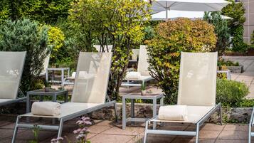 Outdoor lounge chairs with cushions, a white umbrella, and a variety of greenery in the background.