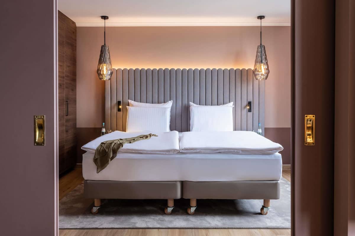 Una cama doble con cabecera gris, dos lámparas de noche y una mesita de noche con una botella.