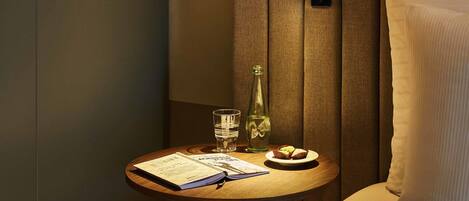 Habitación de hotel ordenada con una pequeña mesita de madera que sostiene un menú, un vaso de agua, una botella de agua y un plato con un muffin.
