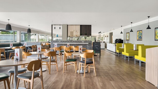 A modern dining area with wooden chairs and tables, a kitchen area, and yellow seating on the right. - Holiday Inn Norwich by IHG (Norwich)