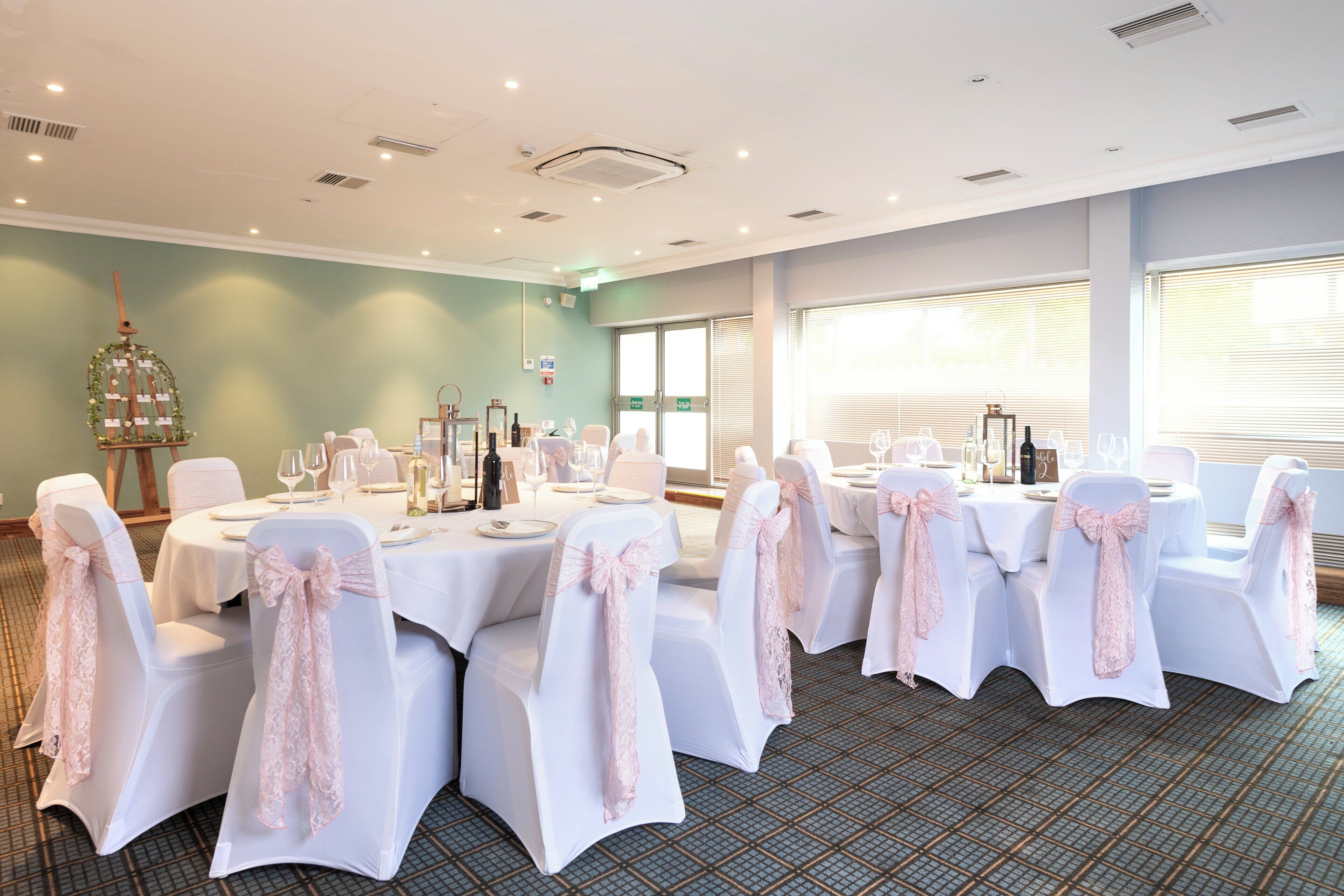 Une salle de conférence avec des tables rondes, des chaises blanches ornées de rubans roses et une cage à oiseaux décorative accrochée au mur.