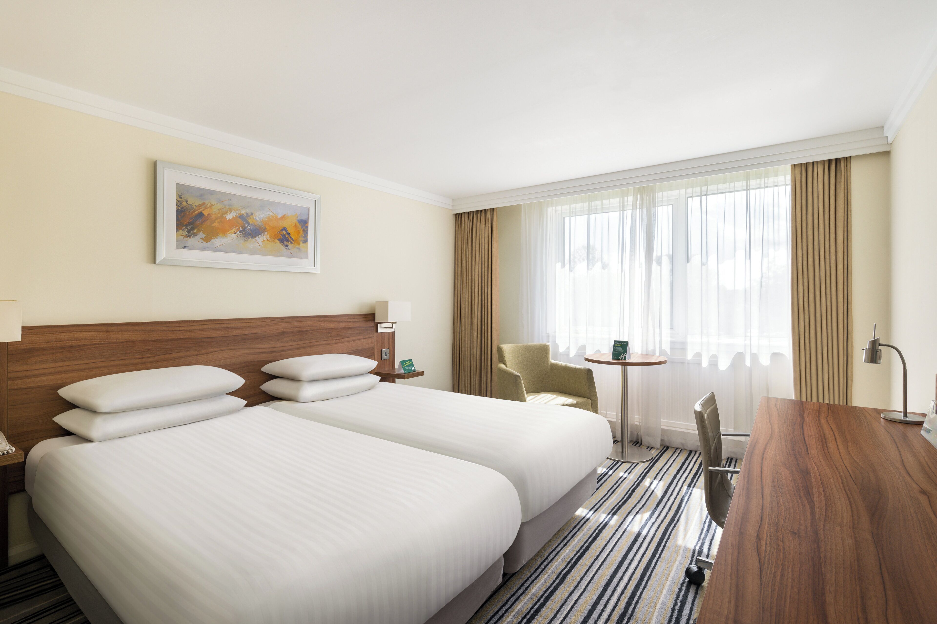 A hotel room with two beds, a desk, a chair, a small table, a lamp, and a painting on the wall.
