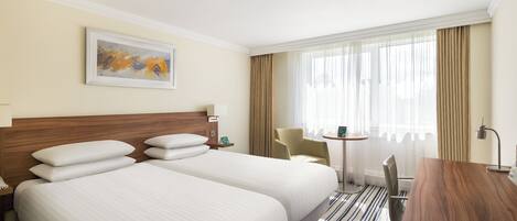 A hotel room with two beds, a desk, a chair, a small table, a lamp, and a painting on the wall.