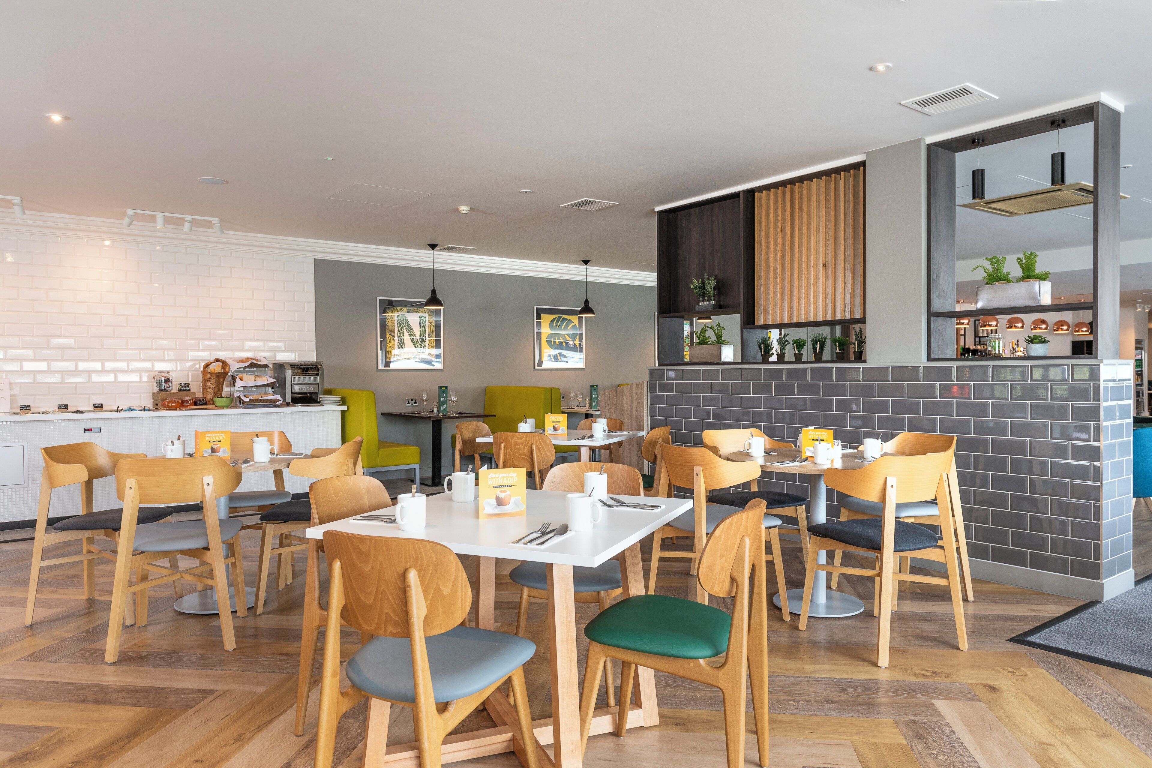 A modern cafe with wooden chairs and tables, a kitchen area, and a brick wall with plants.