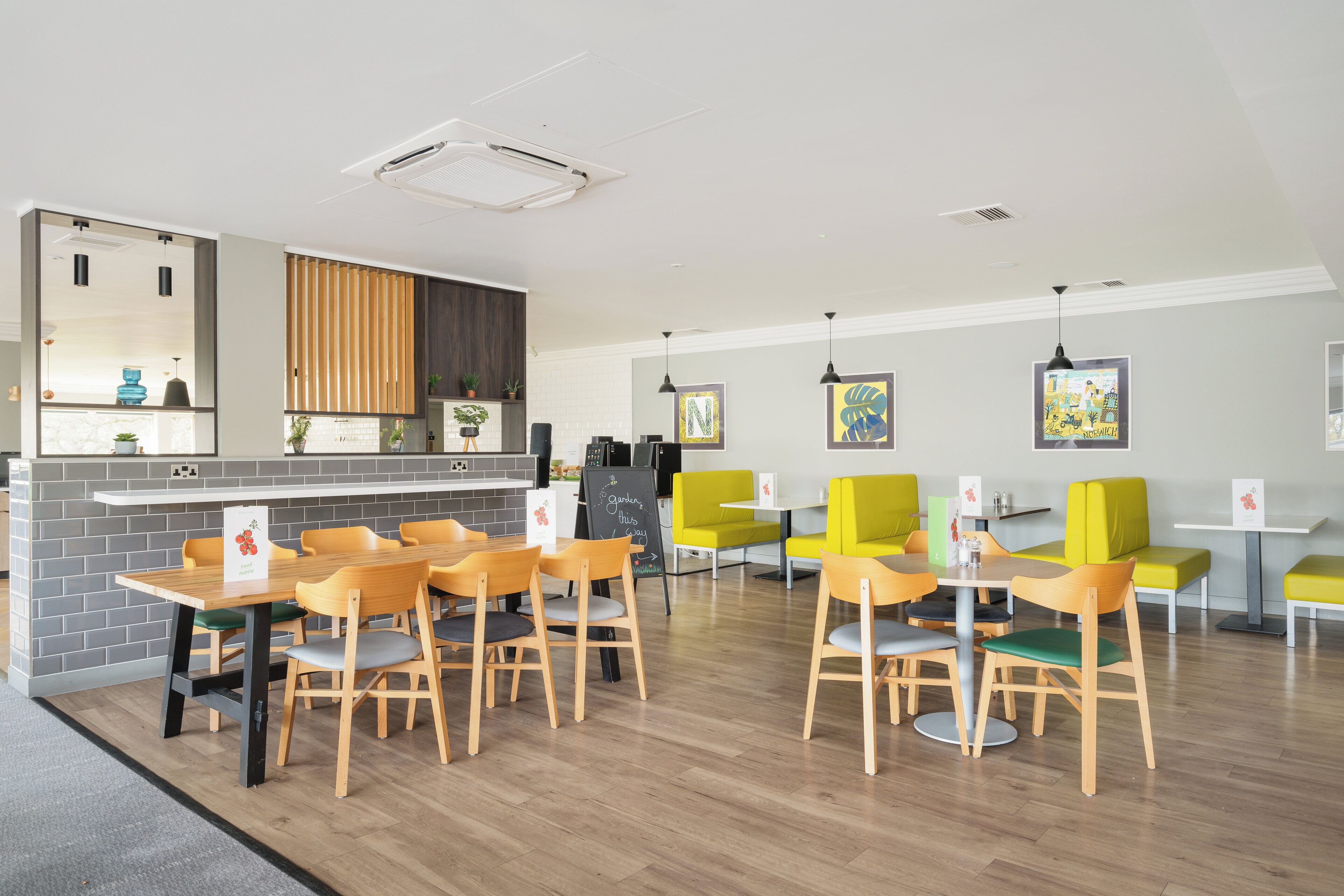 A modern dining area with wooden tables and chairs, a kitchen counter, and a seating area with yellow chairs.