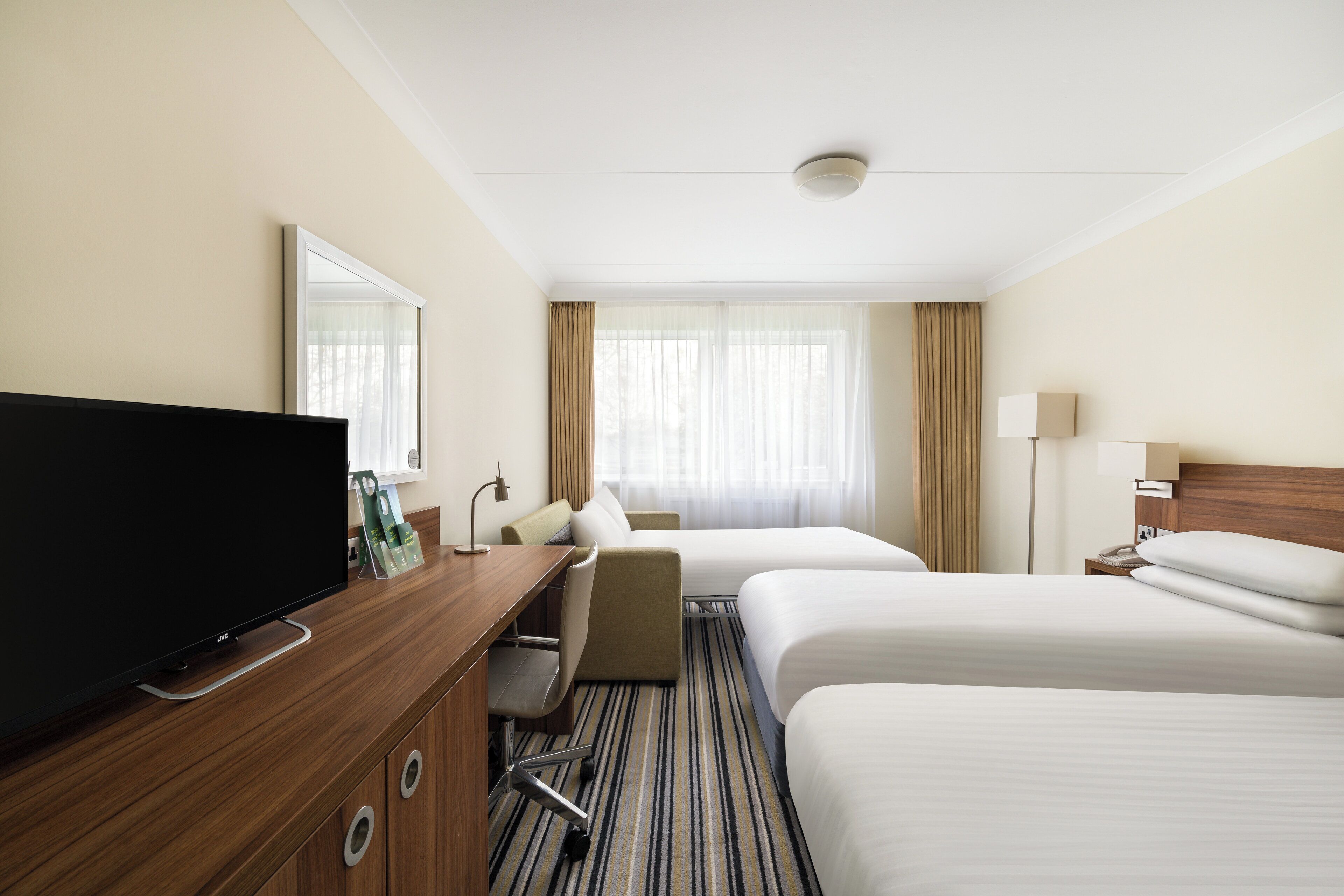 A hotel room with a flat-screen TV, two beds, a desk with a chair, a lamp, and a window with curtains.
