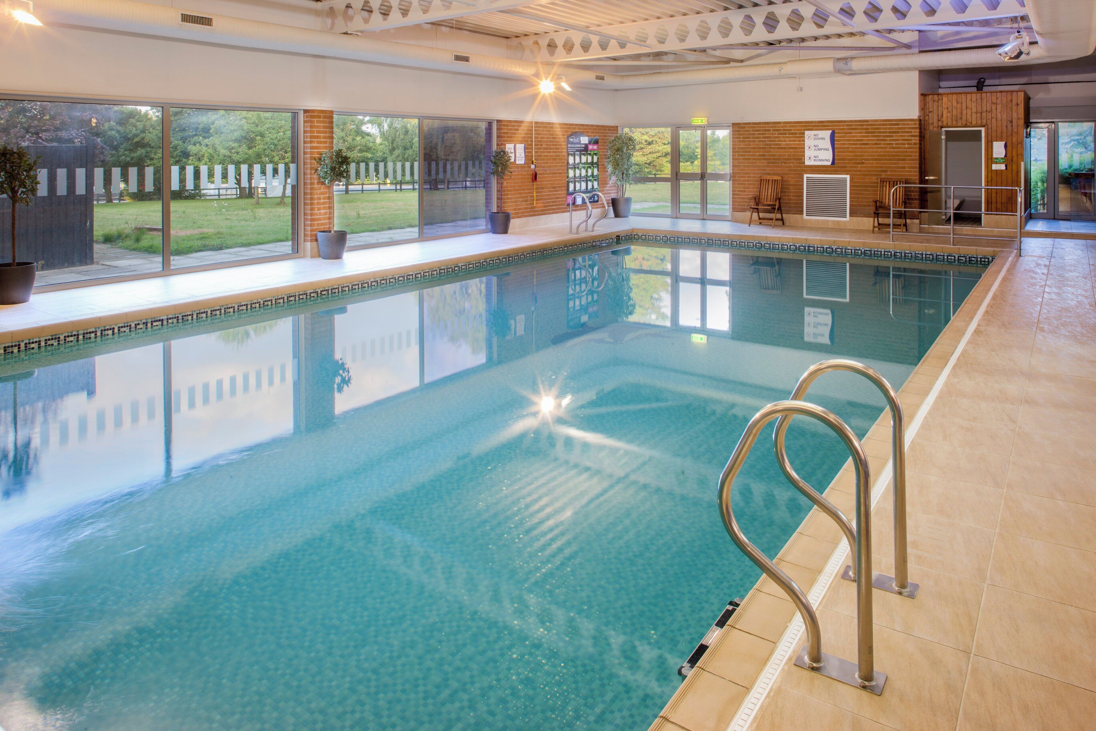 An indoor swimming pool with clear water, a metal ladder, and a view of the outdoors through large windows.
