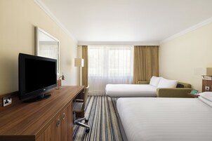 Habitación de hotel con televisión de pantalla plana, escritorio, dos camas, ventana con cortinas y lámpara.