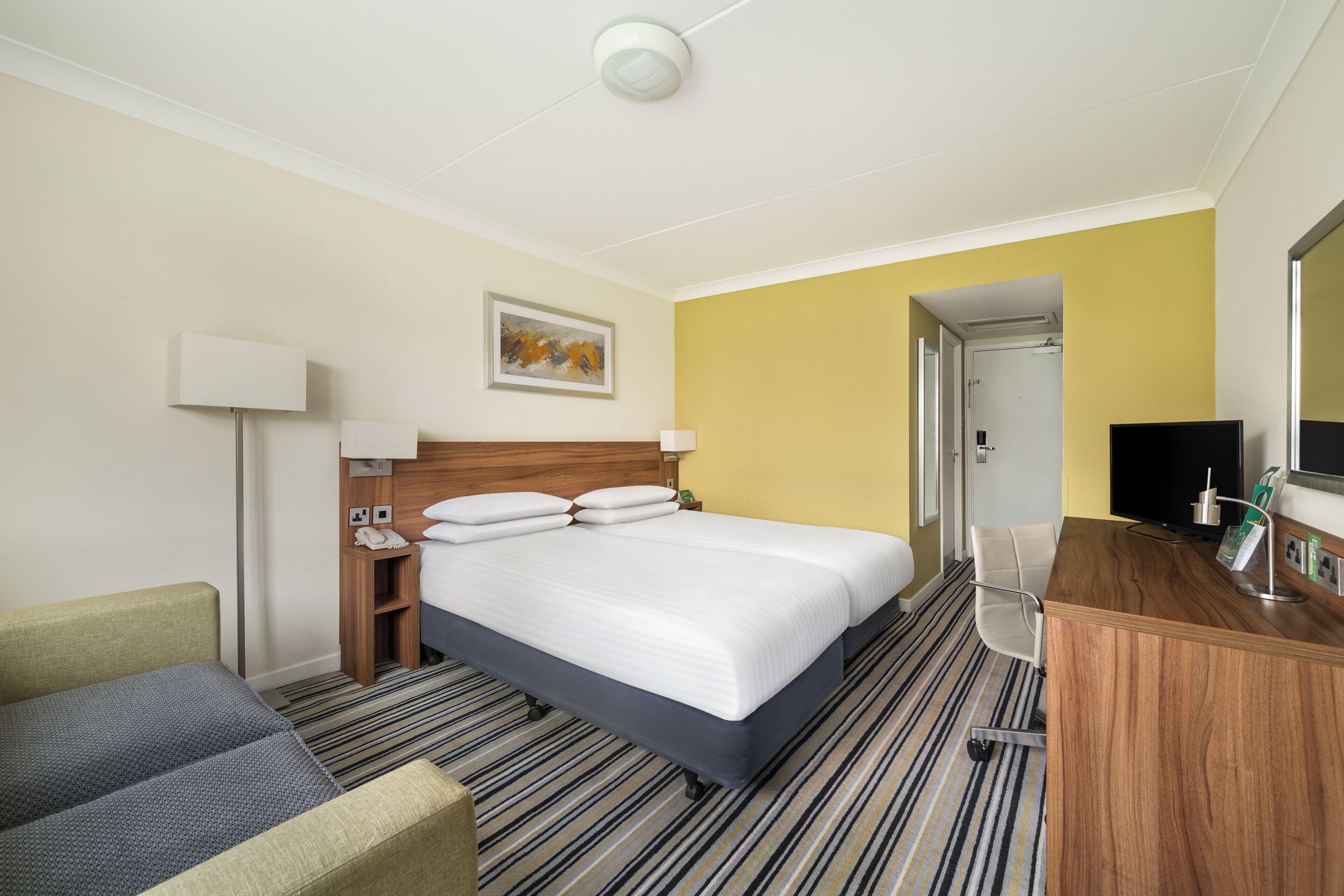 Habitación de hotel con cama, mesitas de noche, escritorio, silla, lámpara, un cuadro y televisor.