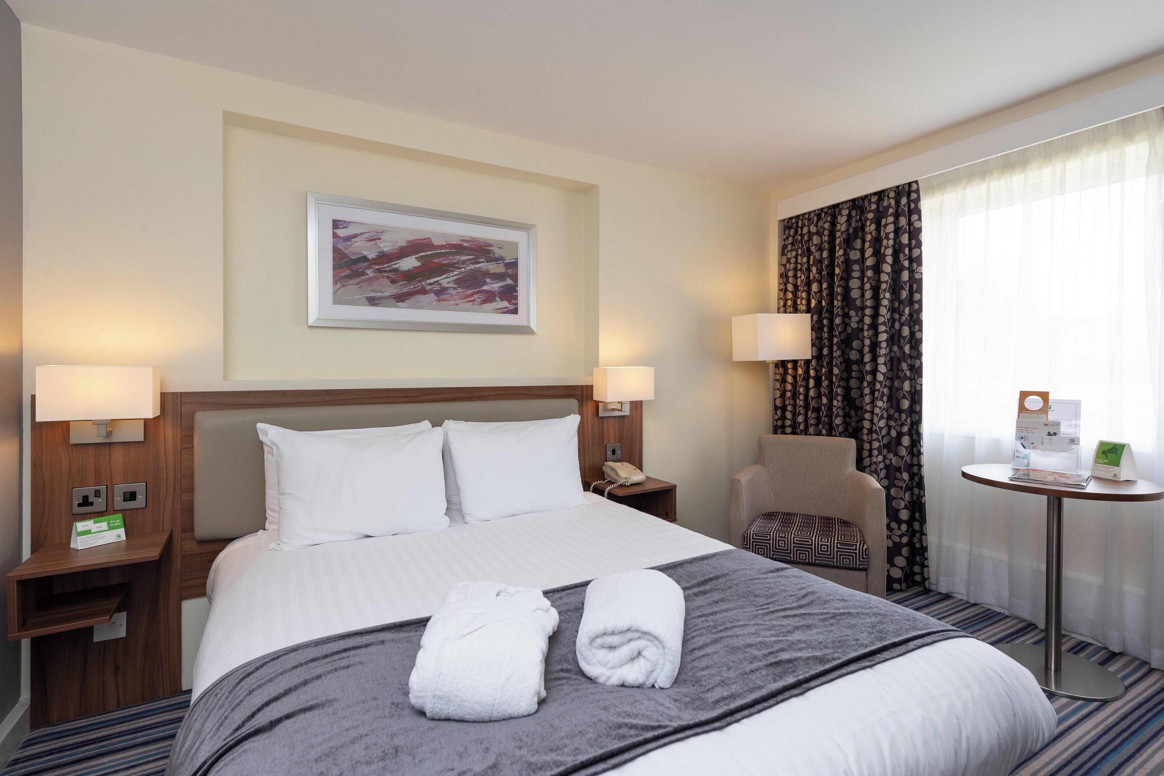 Una habitación de hotel con una cama, dos lámparas de noche, un teléfono, una silla, una mesita y un cuadro en la pared.