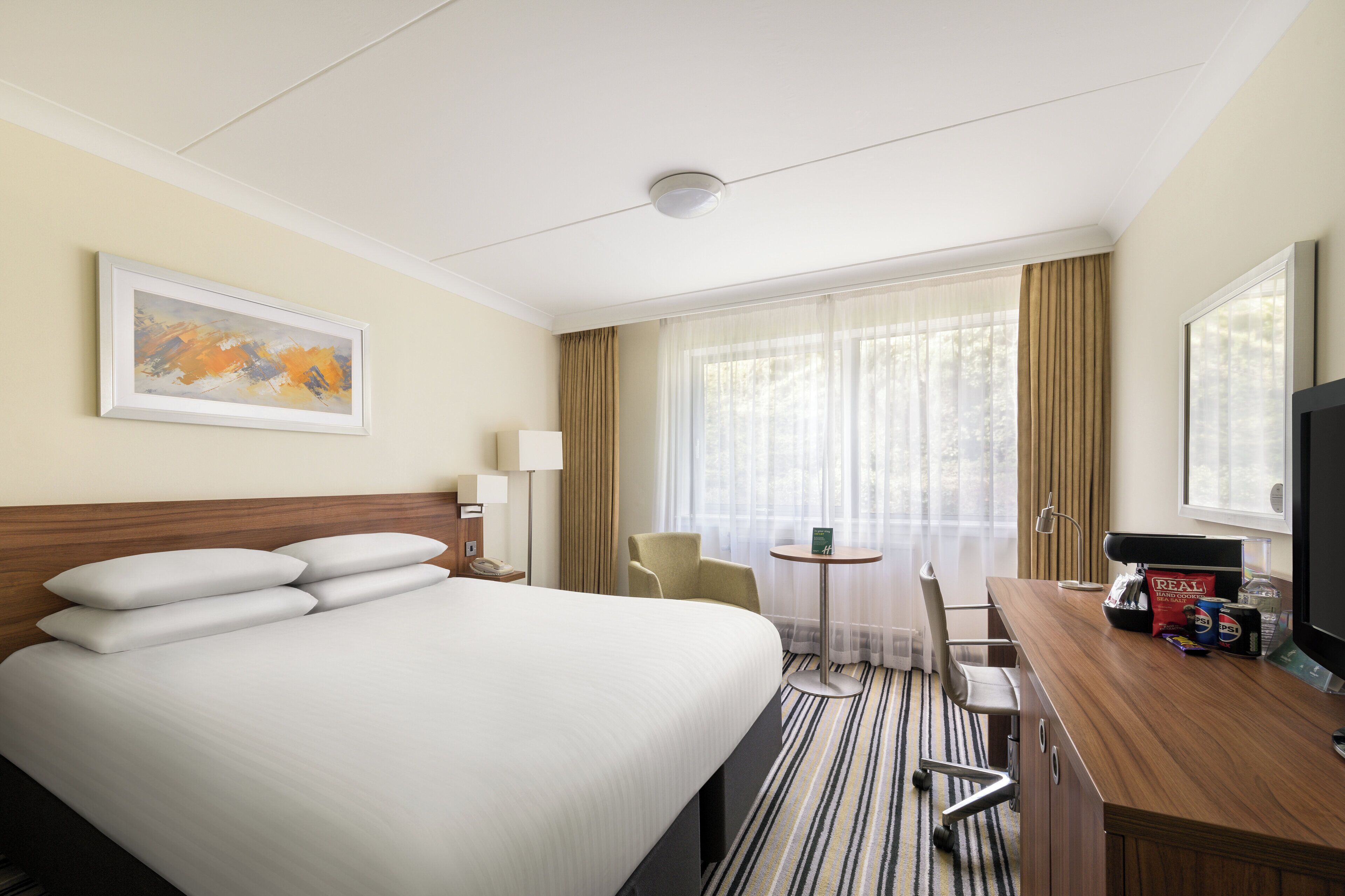 A hotel room with a large bed, a desk with a television, a chair, a small table, and a window with curtains.