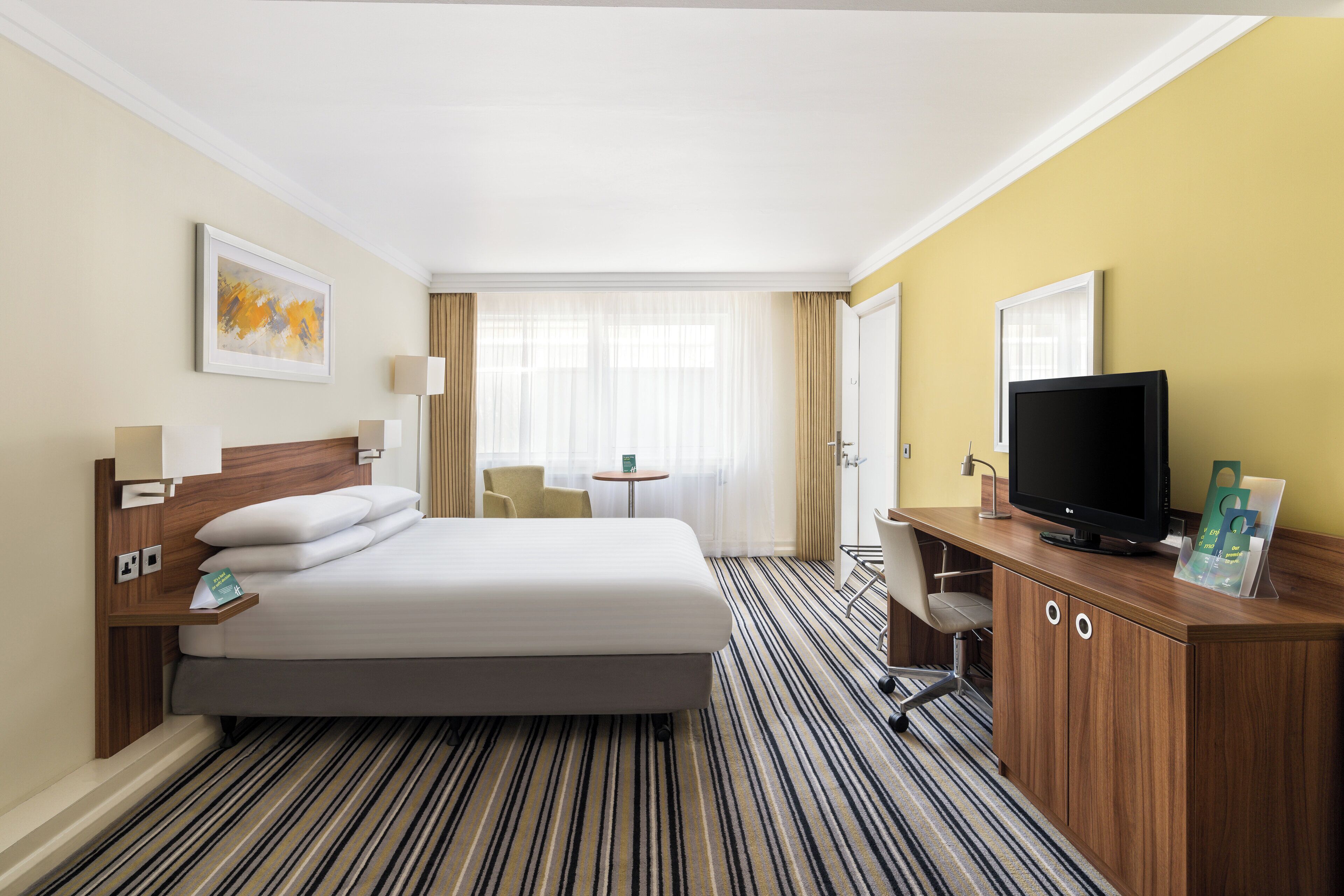 A hotel room with a bed, a desk with a television, a chair, a small table, and a window with curtains.