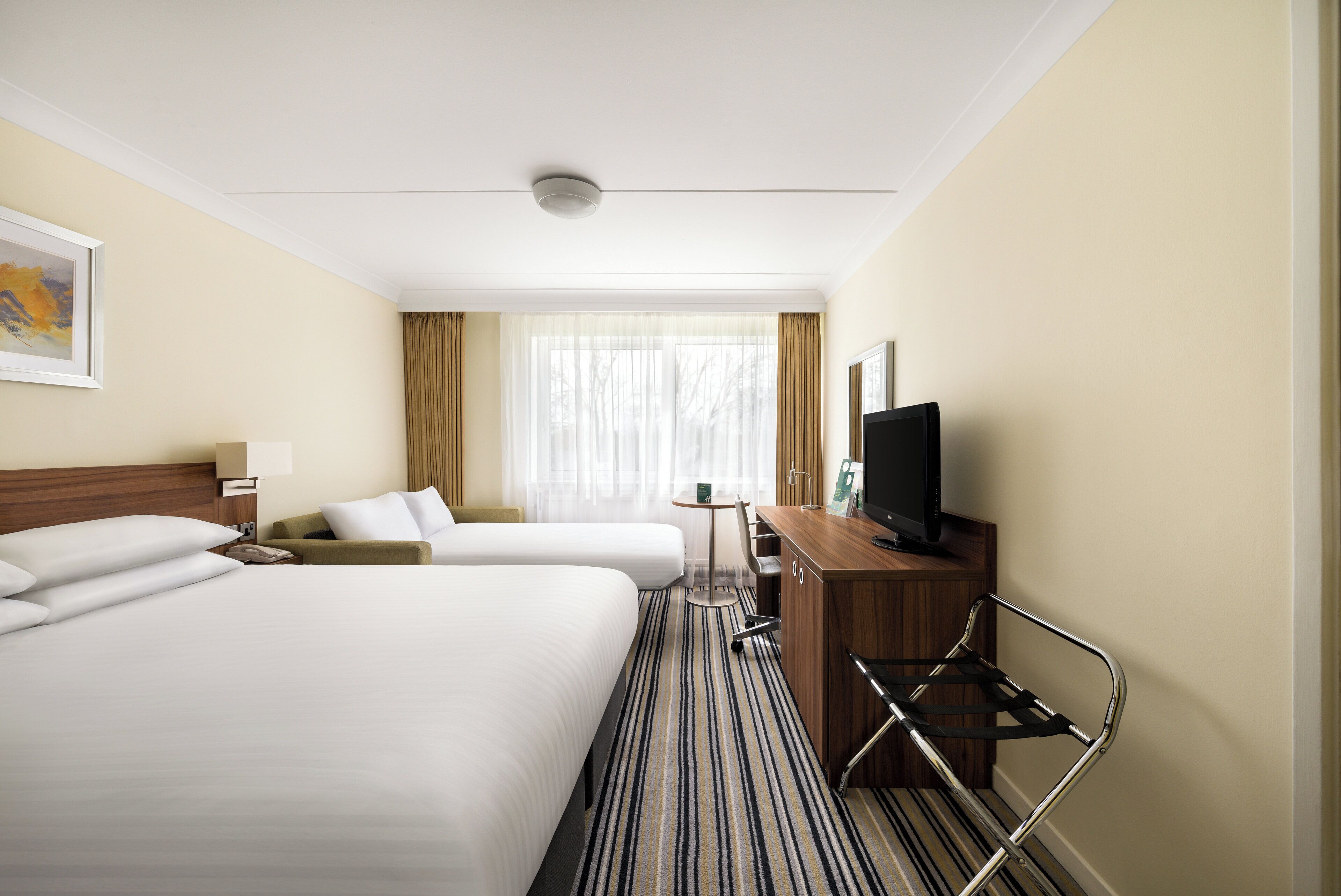 A hotel room with two beds, a desk with a TV, a small table, and a window with curtains.