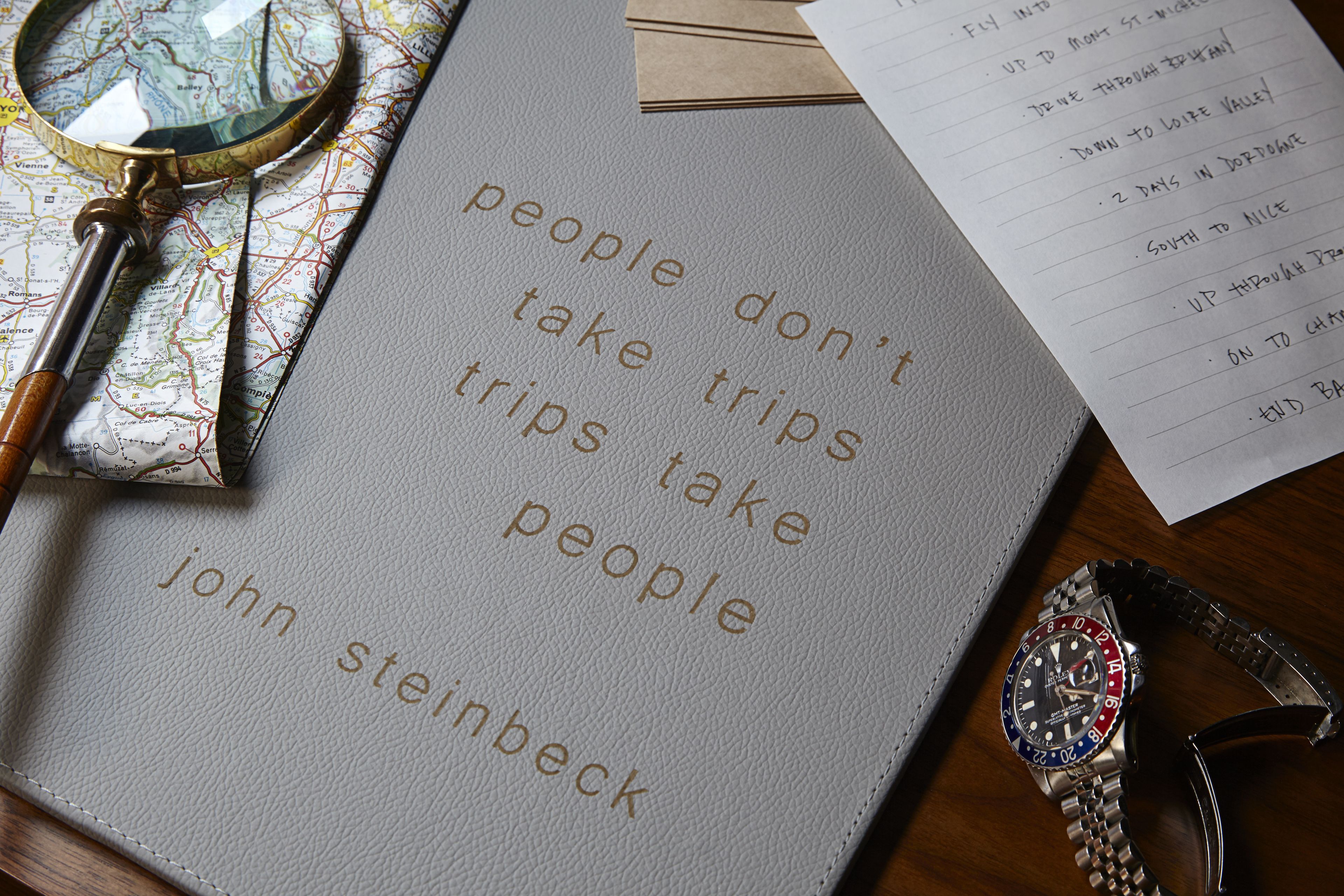 A leather-bound notebook with a quote, a magnifying glass, a wristwatch, and a pair of sunglasses on a wooden surface.