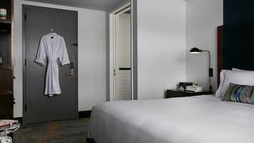 Habitación de hotel con una cama, una mesita de noche con lámpara, un pequeño escritorio, una puerta con una bata colgando y una mesa con un vaso y libros.