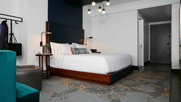 A modern hotel room with a large bed, a nightstand, a desk lamp, a chair, and a door leading to another room.