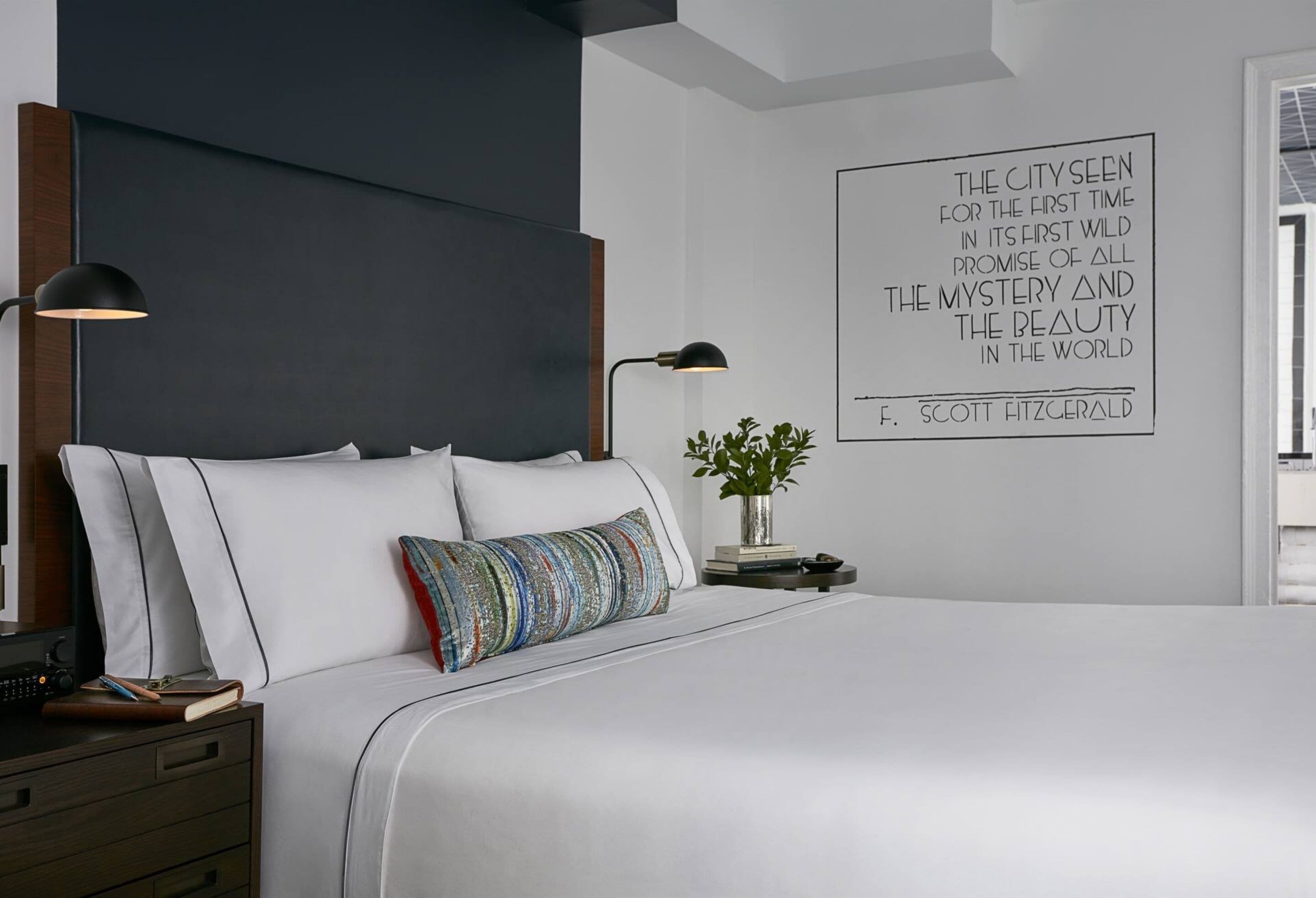 A modern bedroom with a large bed, two bedside lamps, a decorative pillow, a bedside plant, and a motivational quote on the wall.