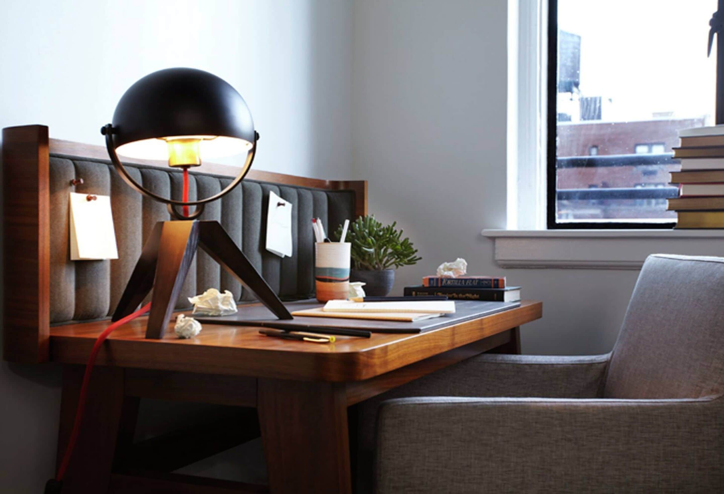 Un escritorio con una lámpara, libros, una planta y una silla en una habitación con ventana.